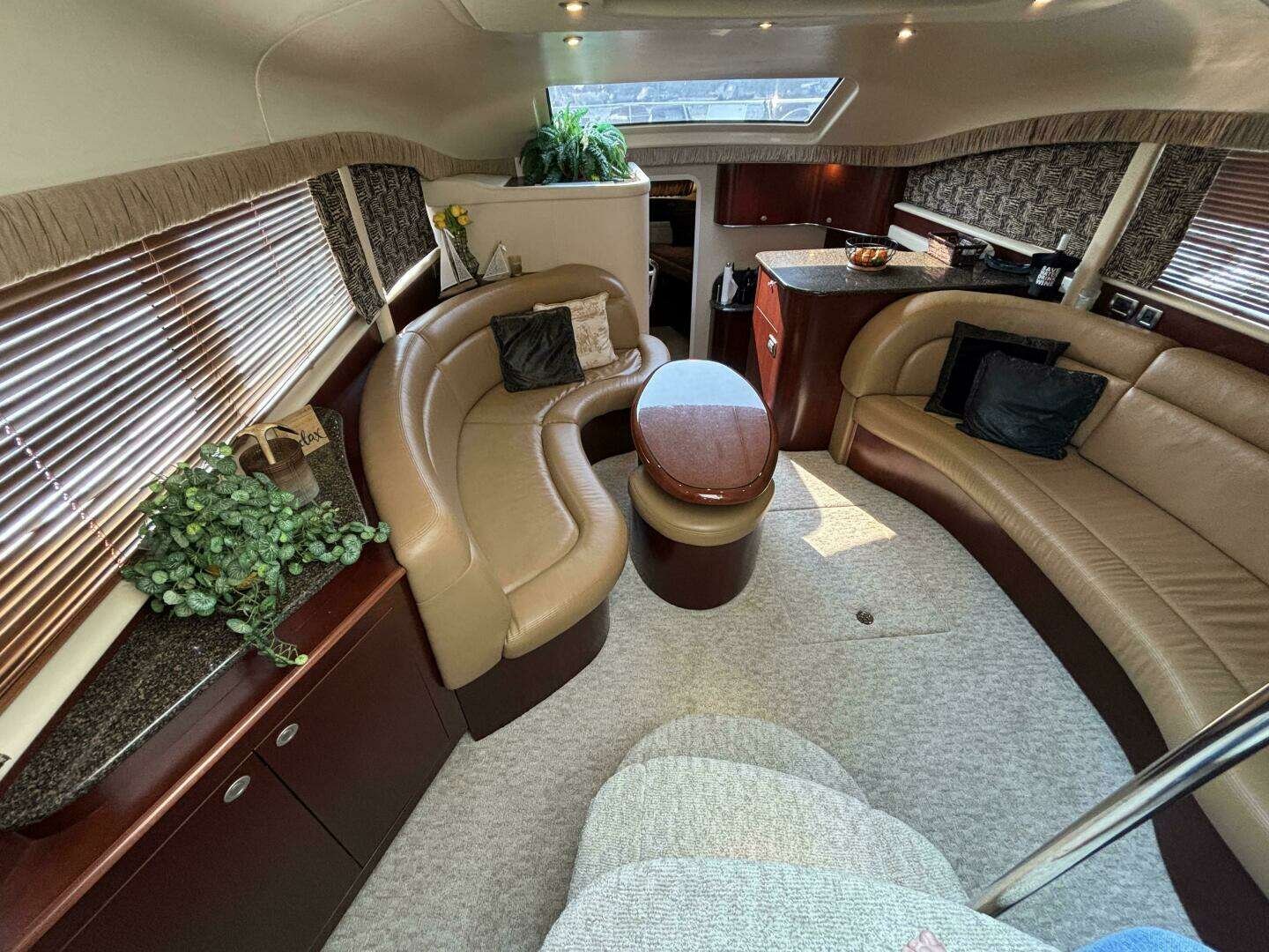 Interior of a Sea Ray yacht, with curved beige leather sofas, wood accents, a small oval wooden table, and windows with blinds and valances. There are decorative pillows, plants, and sunlight coming through a skylight.