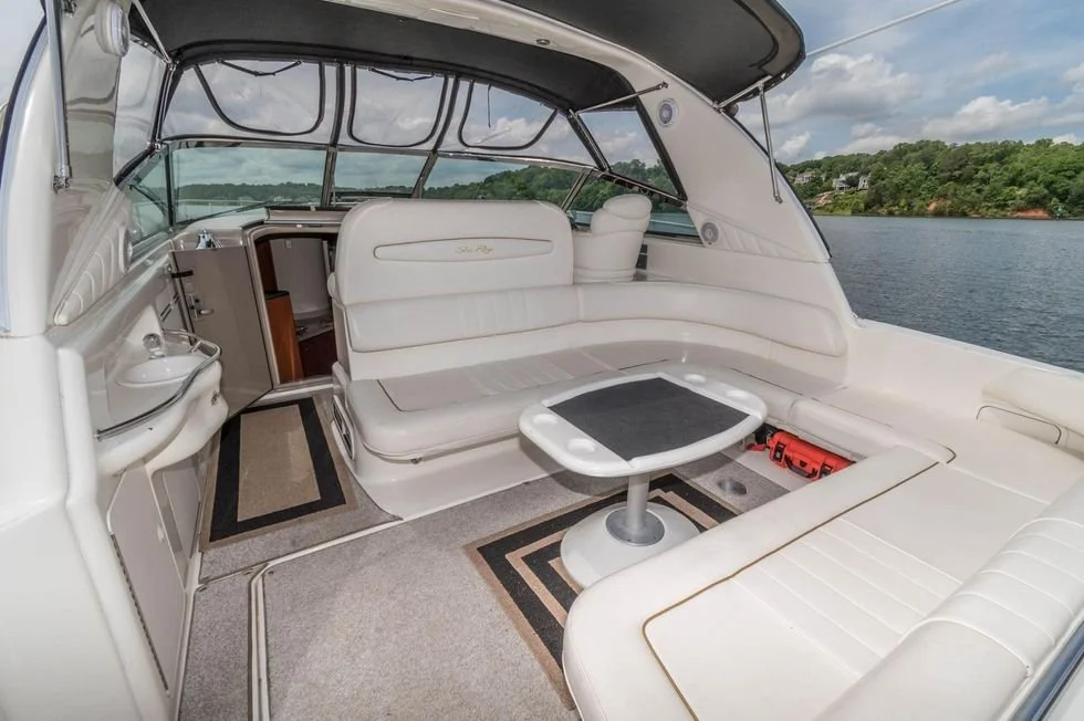 Interior of a yacht with white leather seating, a small table, a kitchenette, and large windows with a view of a lake and trees.