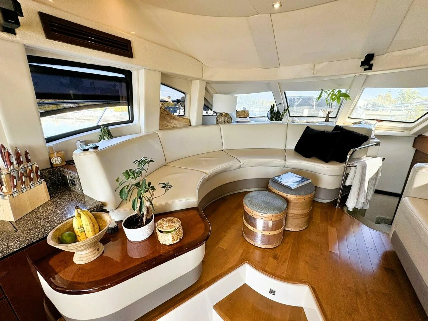 Interior of a yacht with a curved white leather sofa, wooden flooring, and large windows showing water views. The surface has decorative plants, a lamp, books, and small stools. There are black pillows on the sofa and a small table, with various deco
