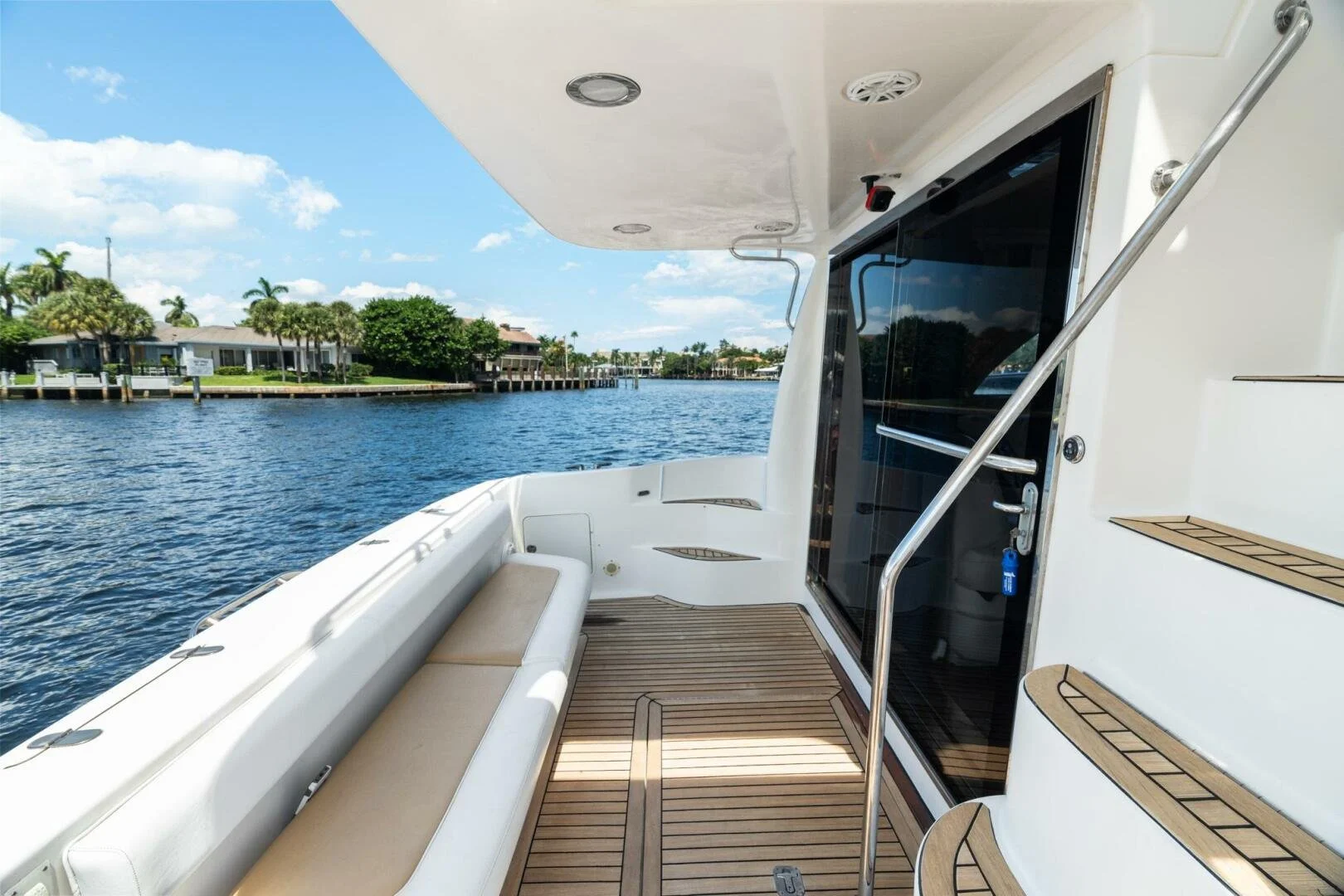 View of boat deck with brown seating and stairs on the right, overlooking a waterway with houses and palm trees under a partly cloudy sky.