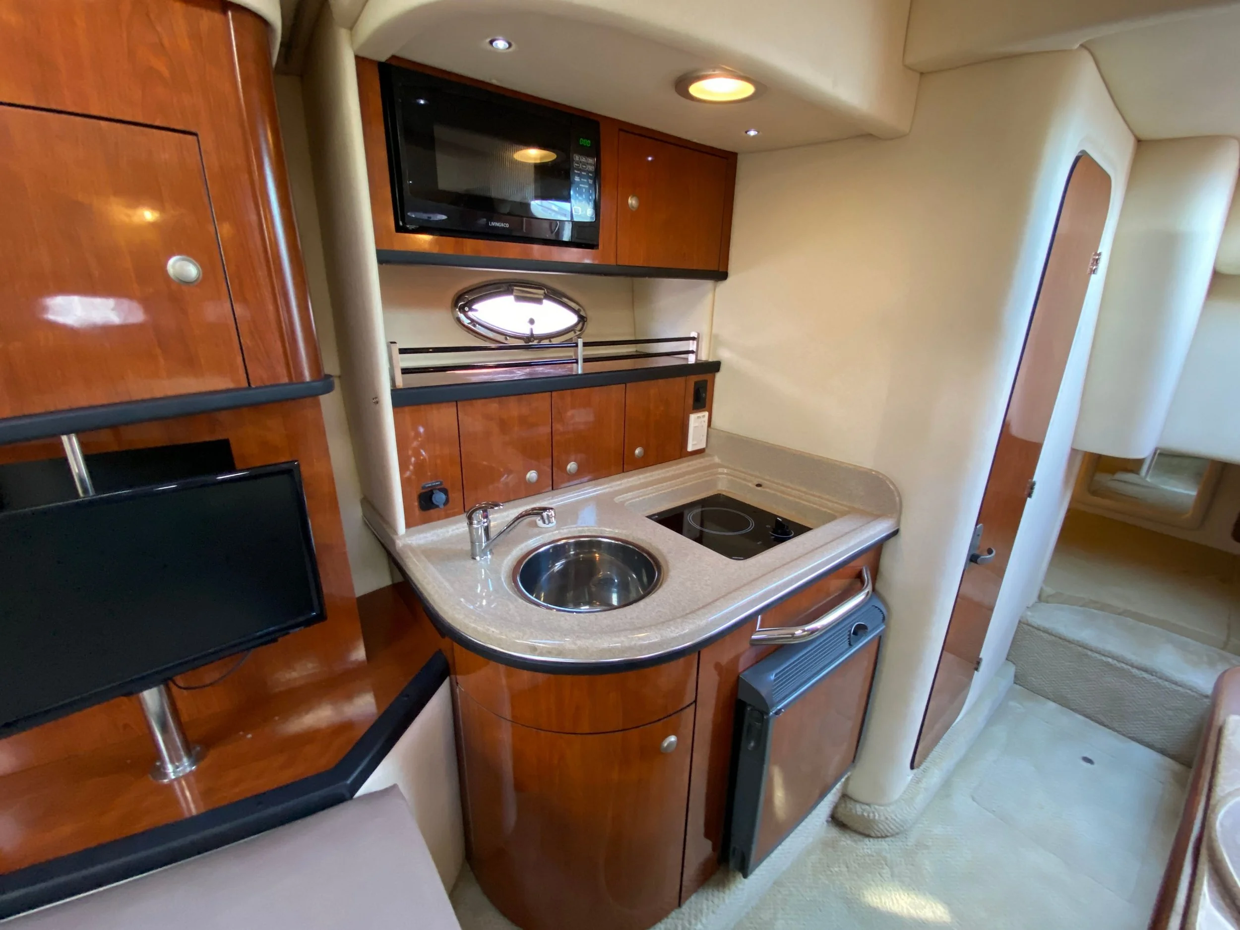 Galley kitchen area with a rounded countertop, built-in sink, microwave, and electric cooktop within a yacht or boat interior, featuring warm wood cabinetry and cream-colored walls.