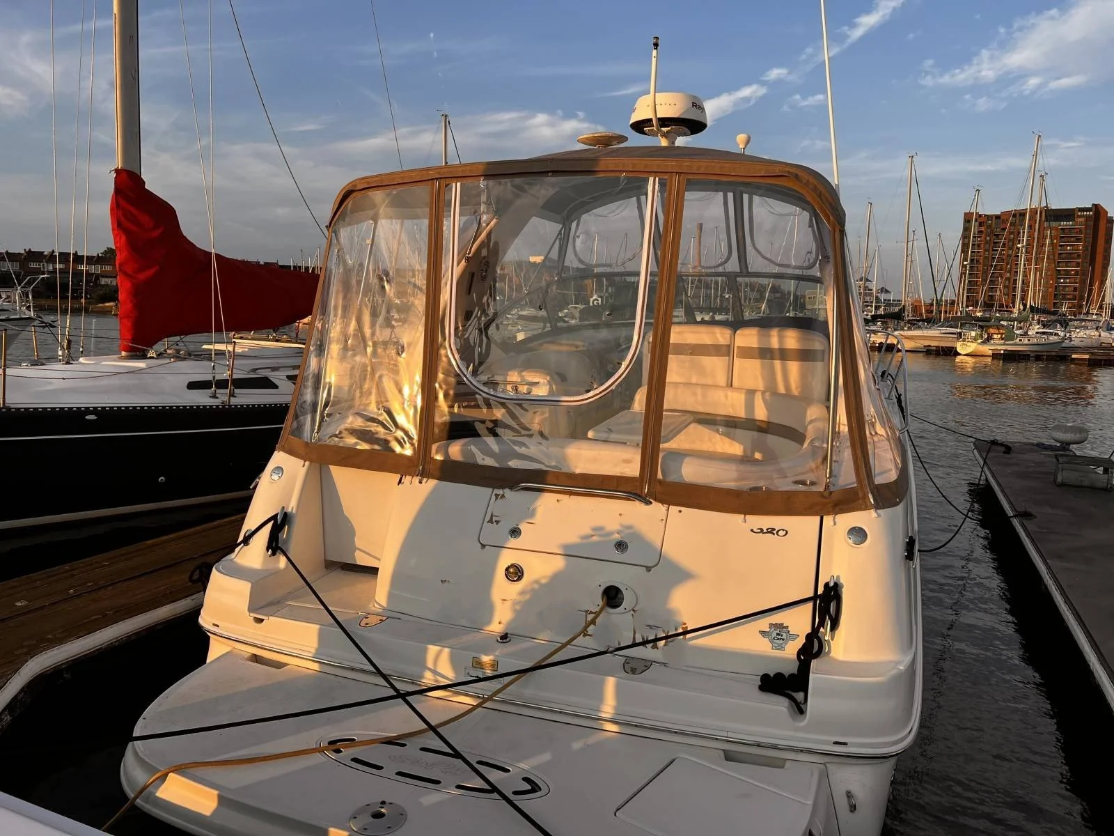 A white boat docked at a marina during sunset with a clear view of its enclosed cabin and seating area, other boats, and a cityscape in the background.