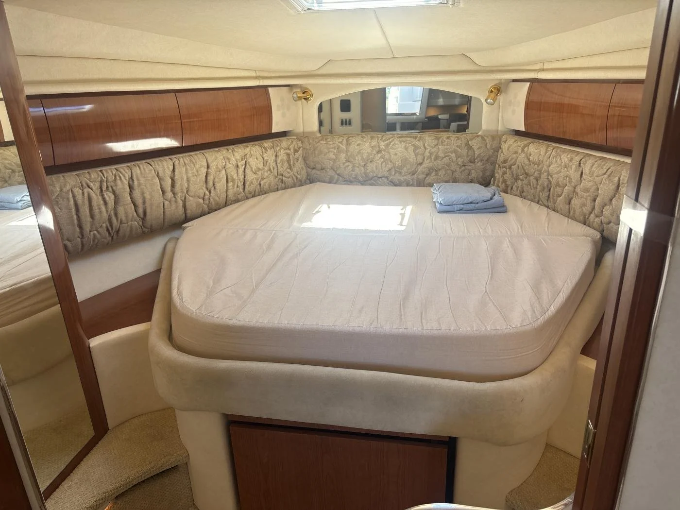 Sea Ray yacht's large, rounded bed with beige bedding and two folded blue towels, surrounded by wood and beige walls and a window with sunlight partially shining on the bed.