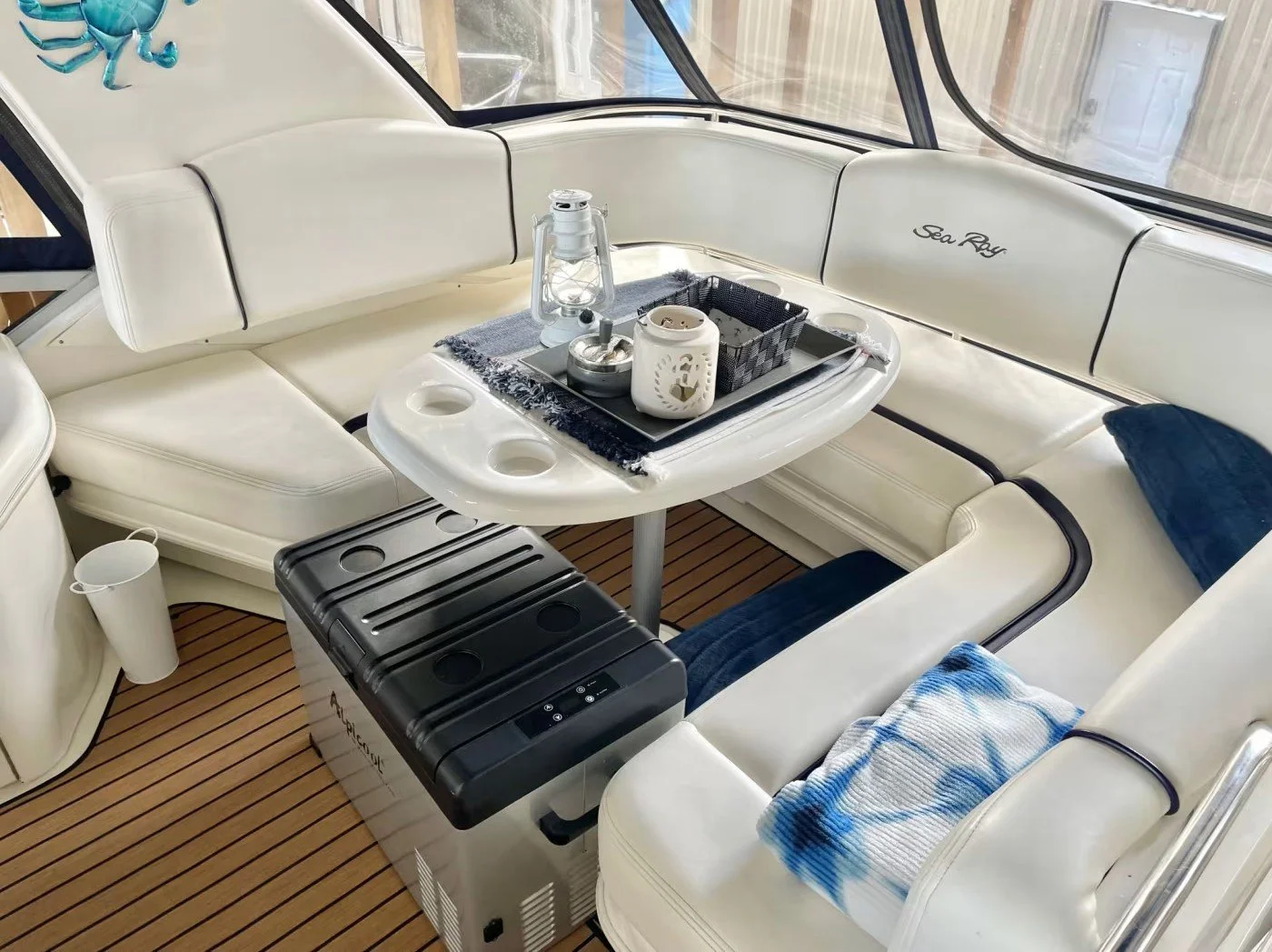 Inside a Sea Ray yacht with white cushioned seating and a table set with a lantern, candle, and other items. The floor has faux wooden planks, and there is a small black refrigerator or cooler nearby.
