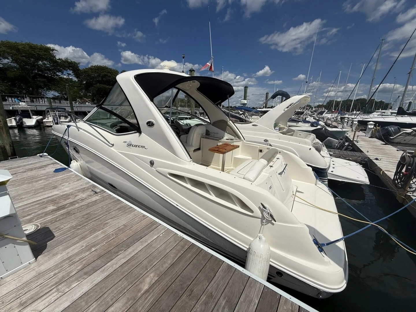 A white yacht docked at a marina pier, surrounded by other boats and sailboats. A sunny summer day with blue sky and scattered clouds in the background. The boat is a Sea Ray 290 Sundancer.