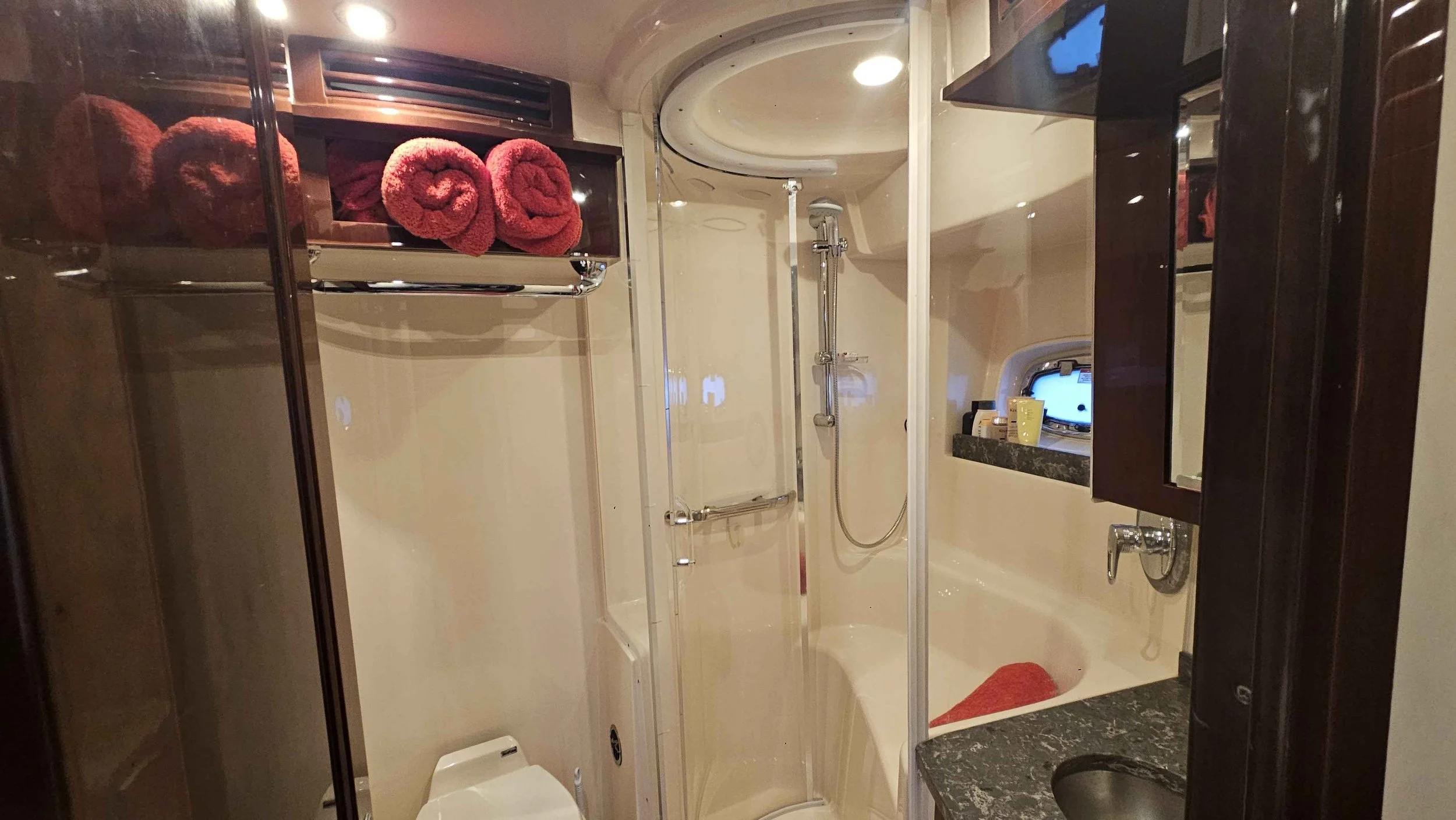 A boat's bathroom with a shower enclosure, a small sink with a granite countertop, a toilet, and a mirror cabinet. Red rolled towels are on a shelf above the toilet, and a small window with a black frame is on the wall. Sea Ray yacht.