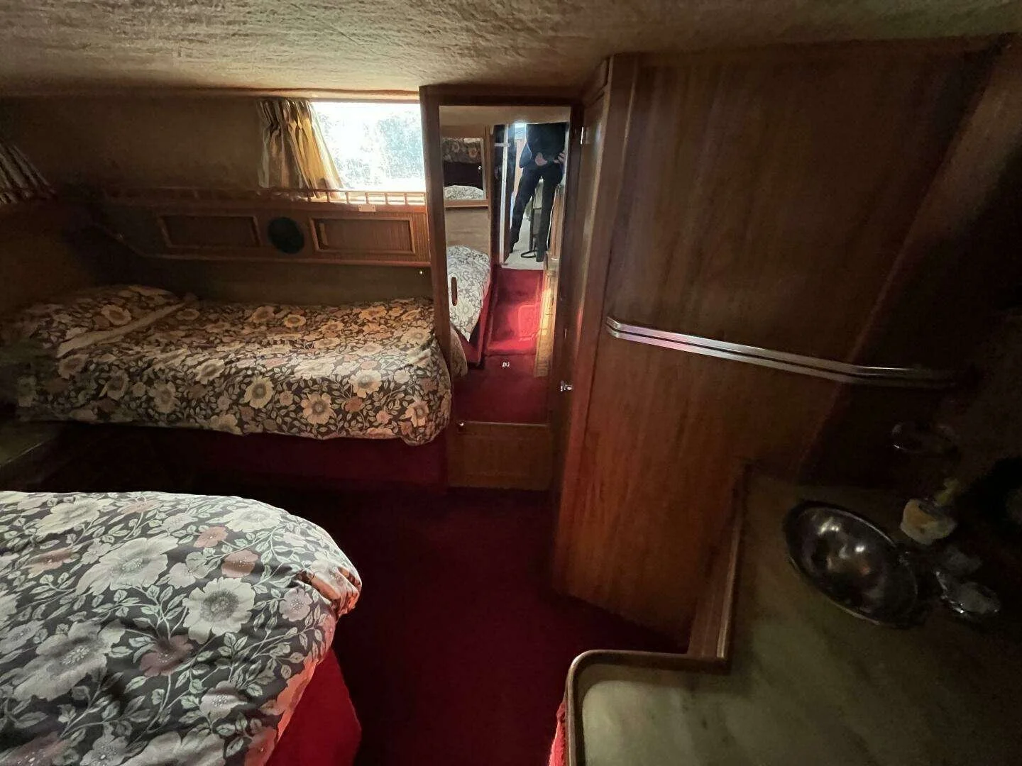 Interior of a vintage Sea Ray yacht, with two beds, wooden paneling, a window with curtains, and a man standing in the doorway taking a photo.