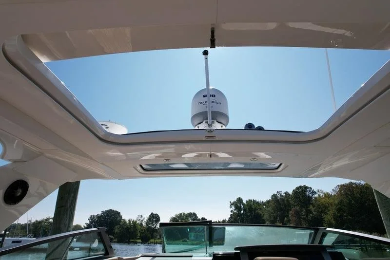 View through the open sunroof of a boat showing a radar and antennas against a clear blue sky, with trees and water in the background.