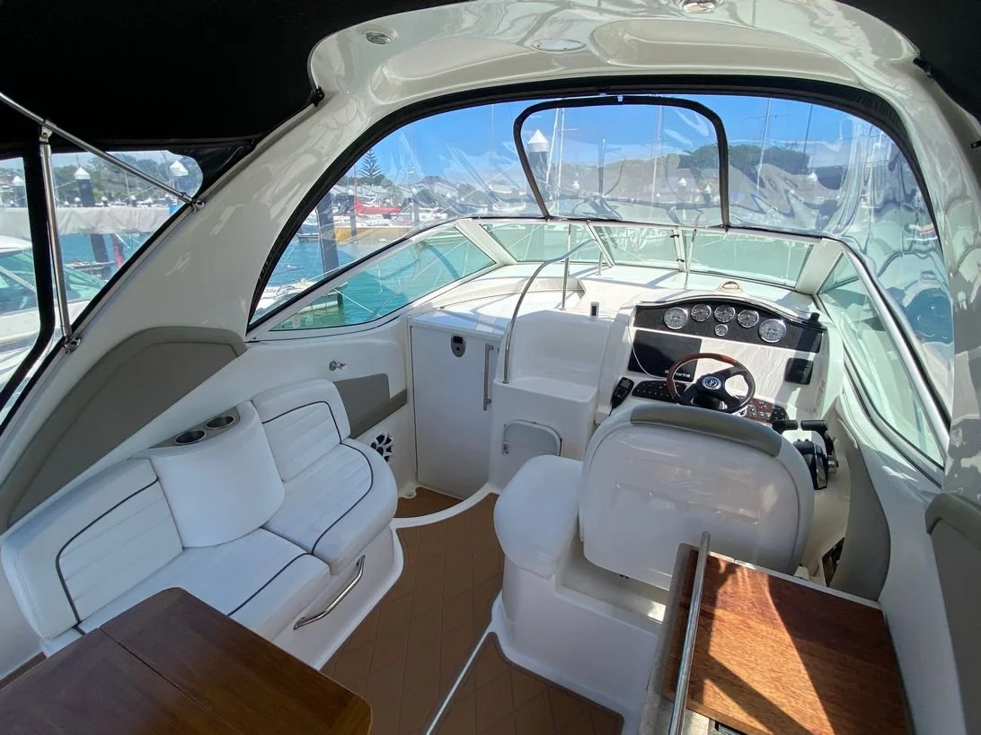 Inside view of a boat cockpit with white leather seating, wooden accents, and a steering wheel, with a marina visible through the windows.