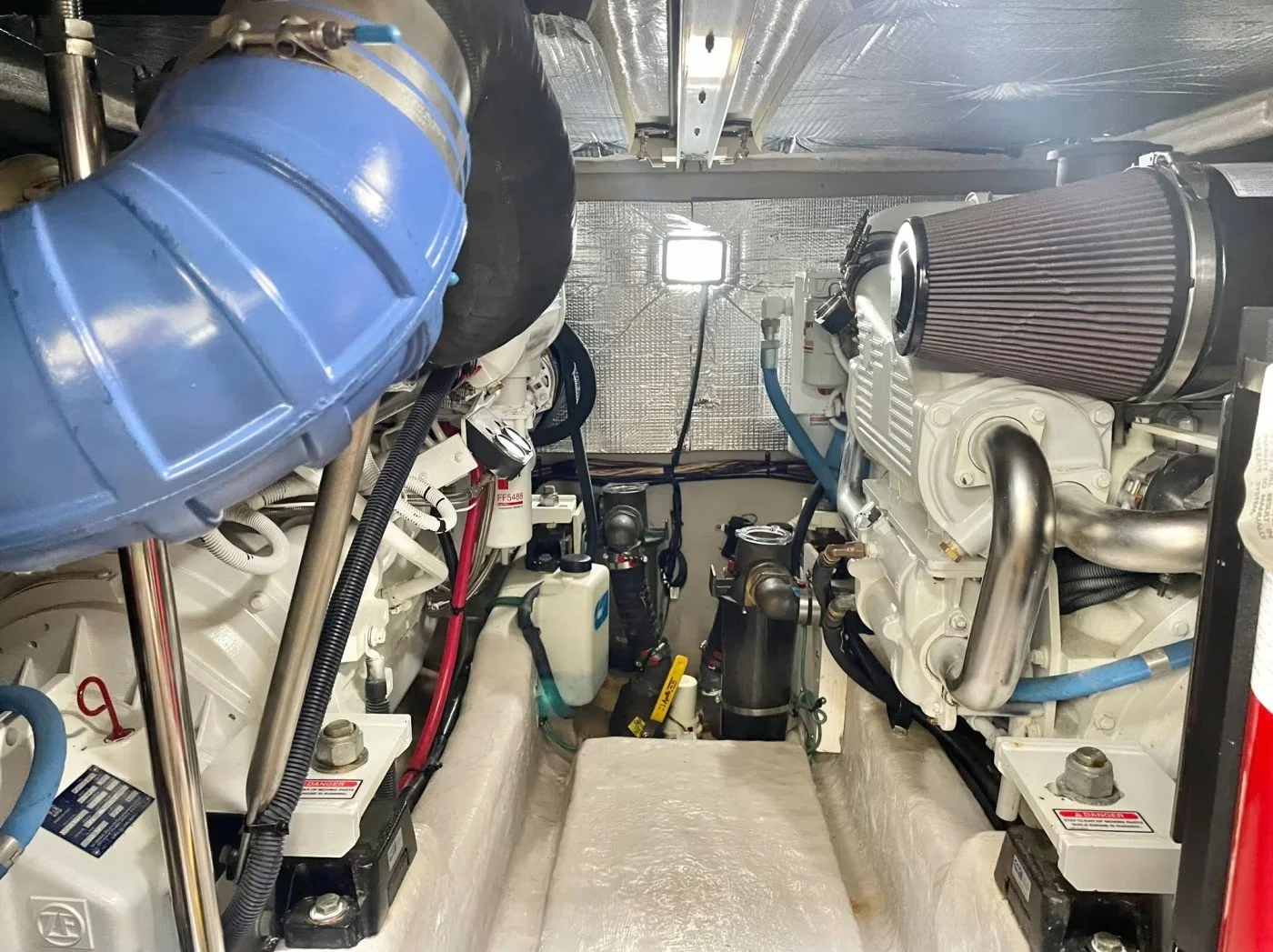 View of a boat engine room showing two large white engines, a blue filter, various hoses and wires, and a flexible brown air filter on the right engine, with metallic walls and ceiling.