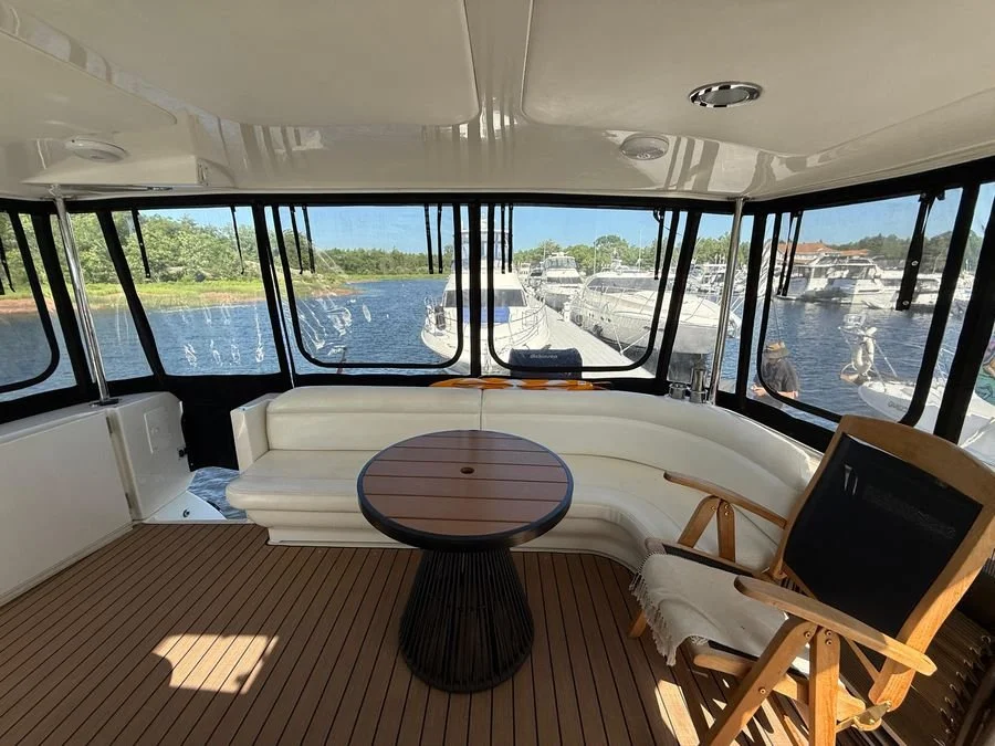 View from inside a boat with large windows showing a marina with boats, a white curved sofa, a round wooden table, and a wooden chair with a towel draped over it.
