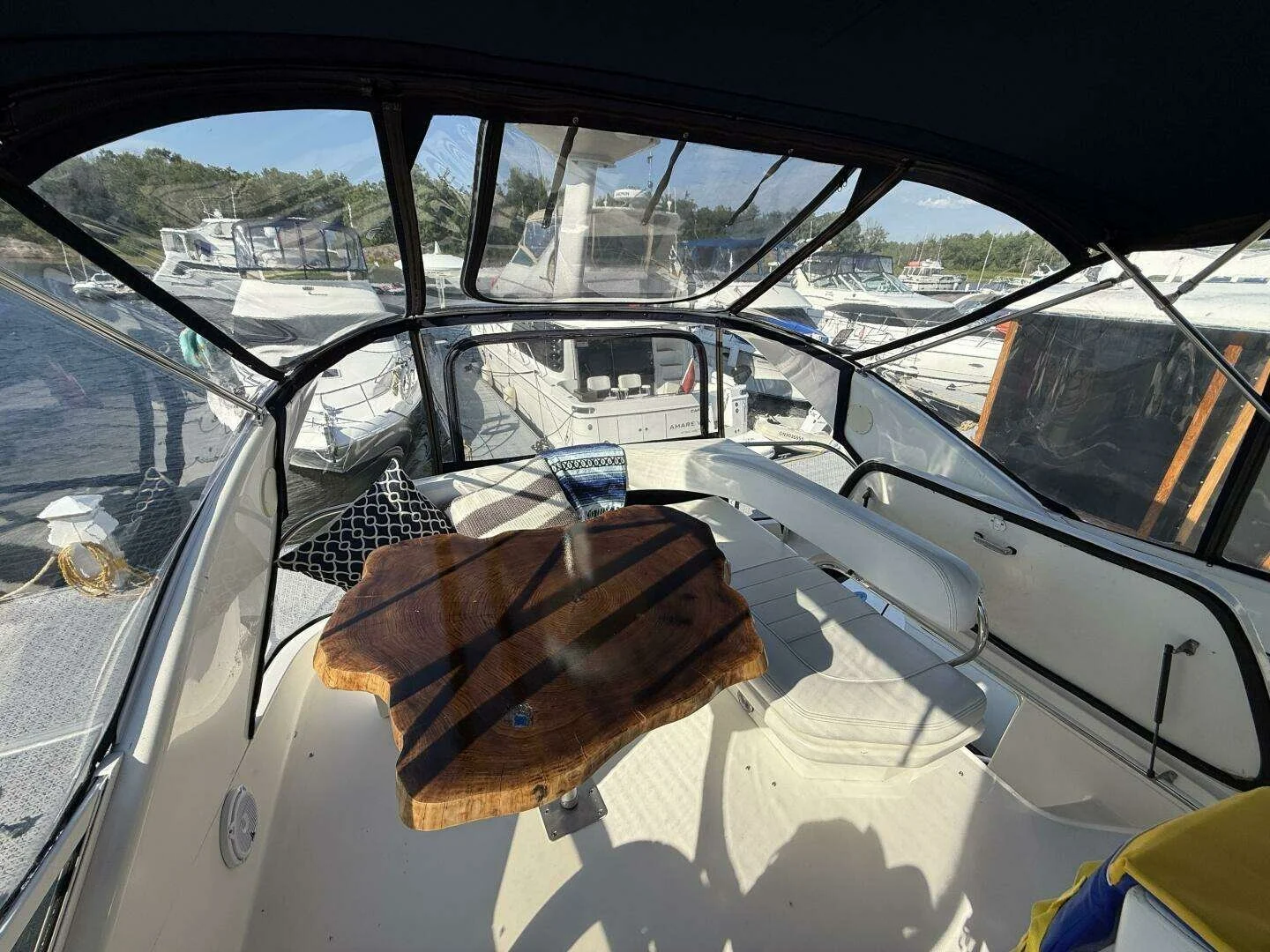 Interior view of a boat with a wooden table, a white cushioned bench, a black and white patterned pillow, and a view of other boats docked at a marina through the windows.