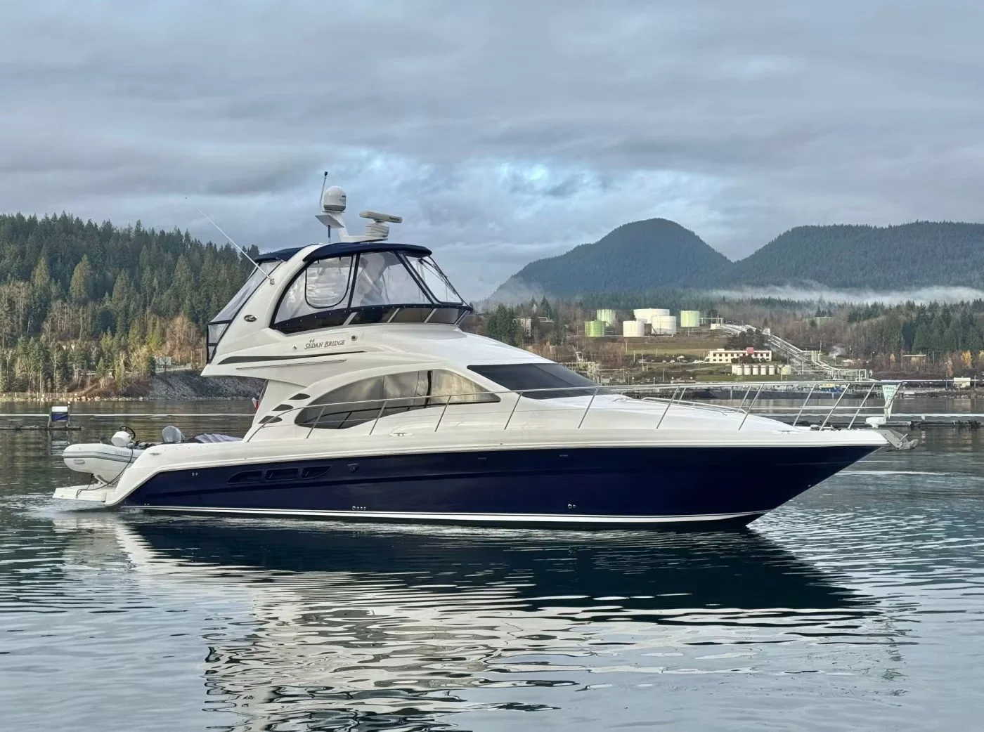 A white yacht with a black stripe is floating on calm water with a backdrop of green hills, and mountains under cloudy sky. Sea Ray 44 Sedan Bridge boat. Flybridge with blue canvas enclosure.
