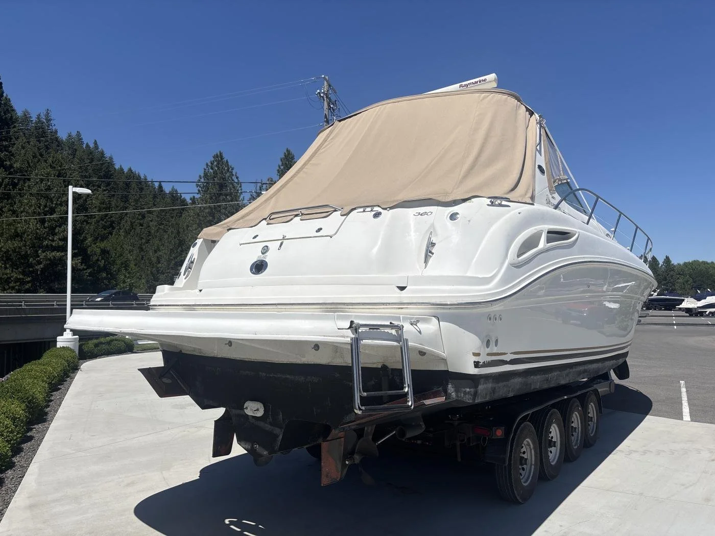 A white yacht on a trailer parked on a paved surface with trees and a clear blue sky in the background.