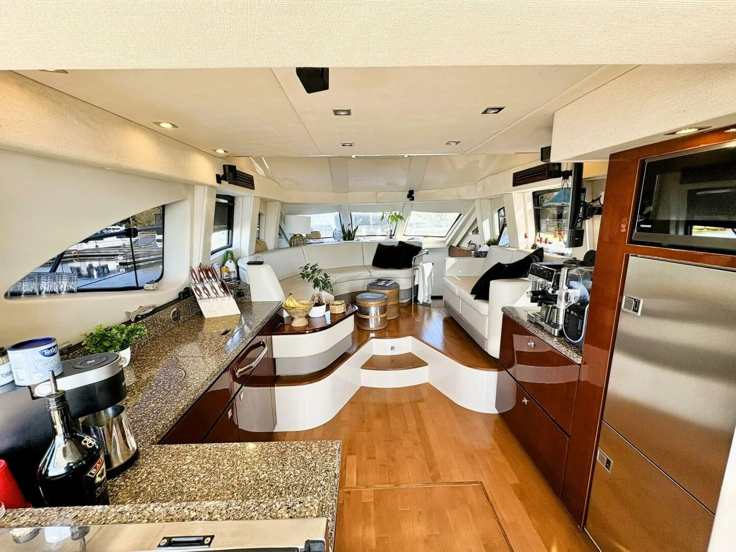 Interior of a yacht with a kitchen area on the left featuring granite countertops, and a living area with white couches, black pillows, and large windows in the background.