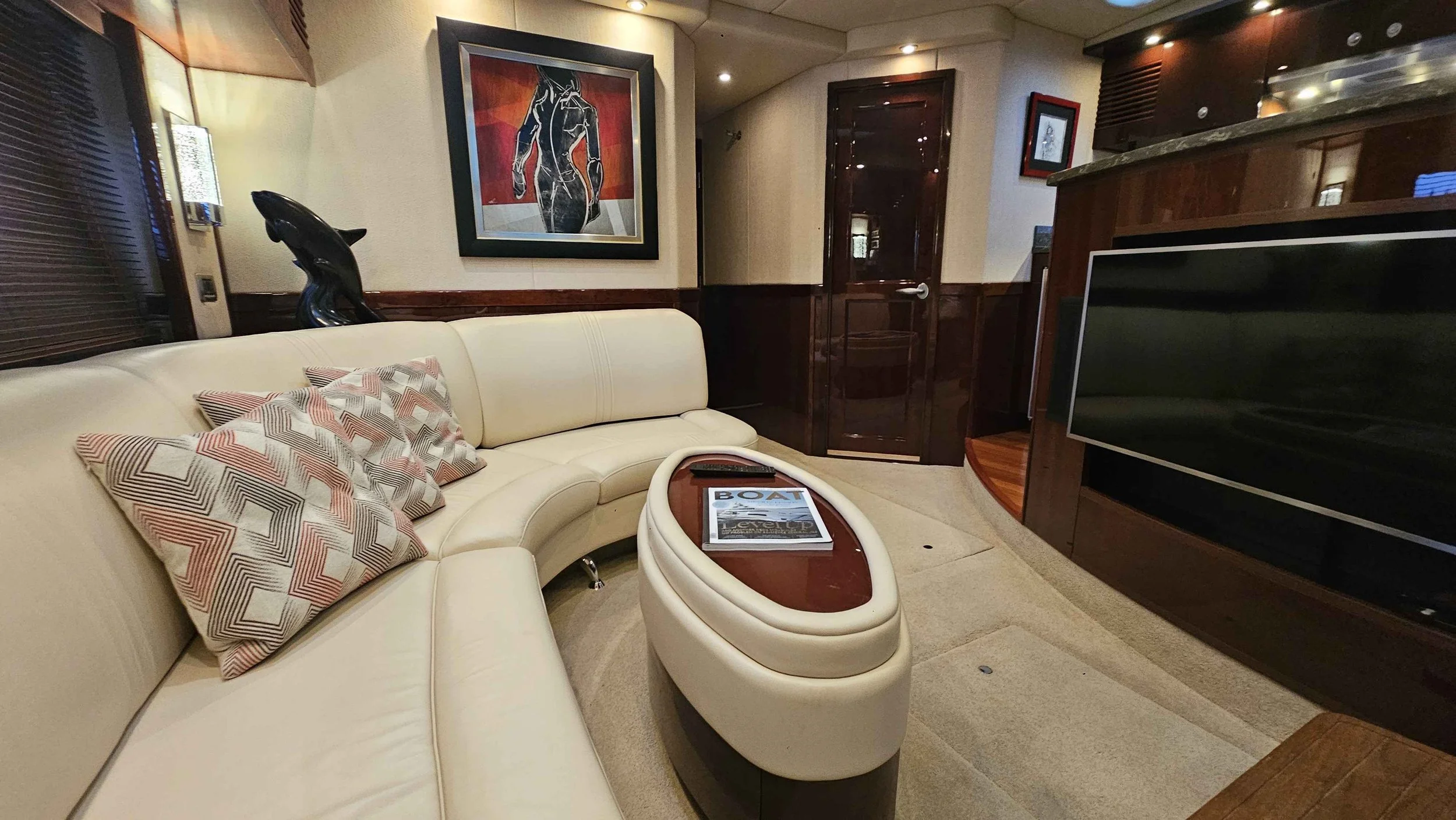 Luxury yacht interior with a cream-colored curved sofa, decorative pillows, a modern oval-shaped coffee table with magazines, a large flat-screen TV, dark wood-paneled walls, abstract art on the wall, and ambient lighting.