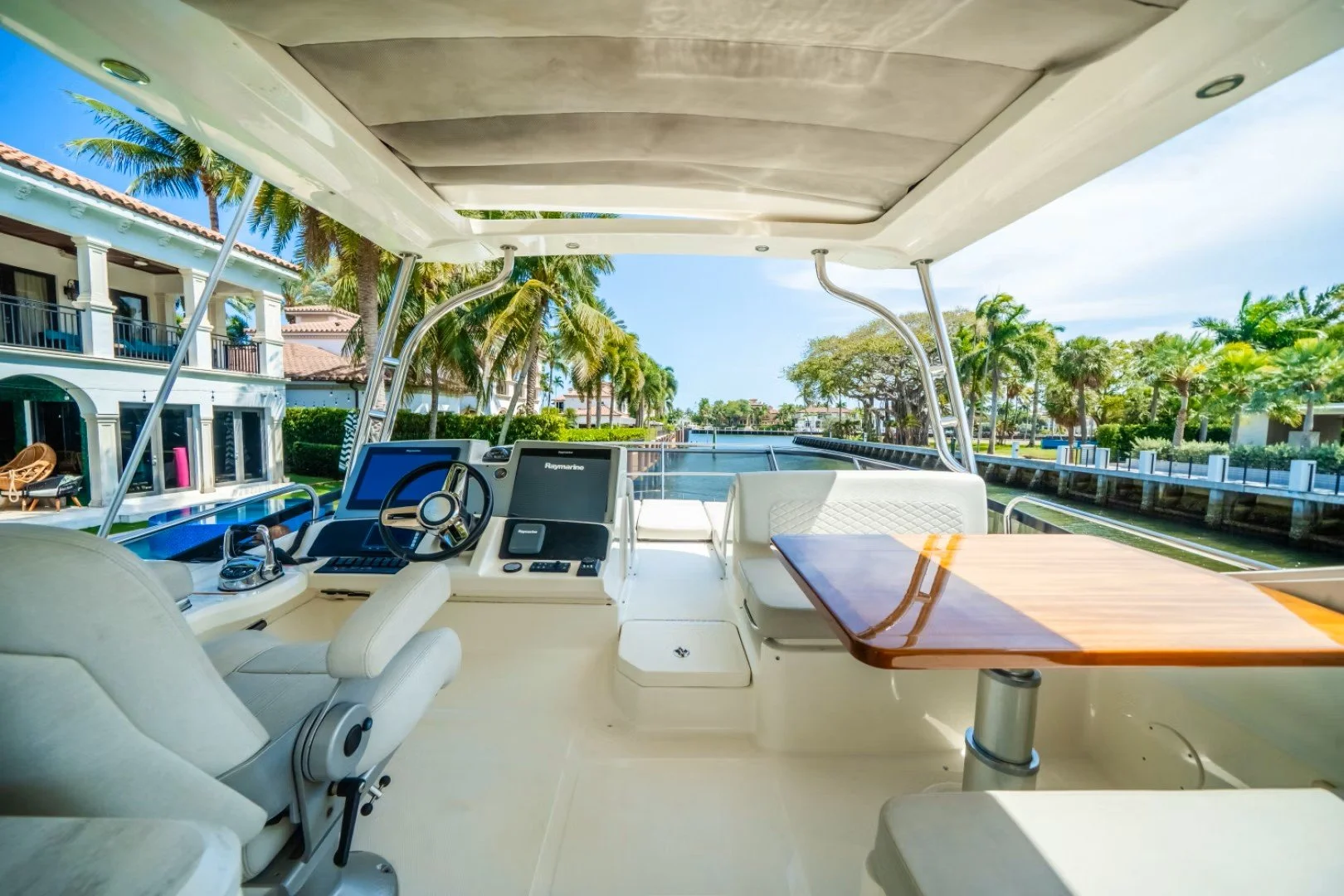 View from the cockpit of a luxury yacht showing the steering wheel, navigation screens, a captain's seat, and a wooden table, with a canal, palm trees, and waterfront houses in the background. Sea Ray boats.