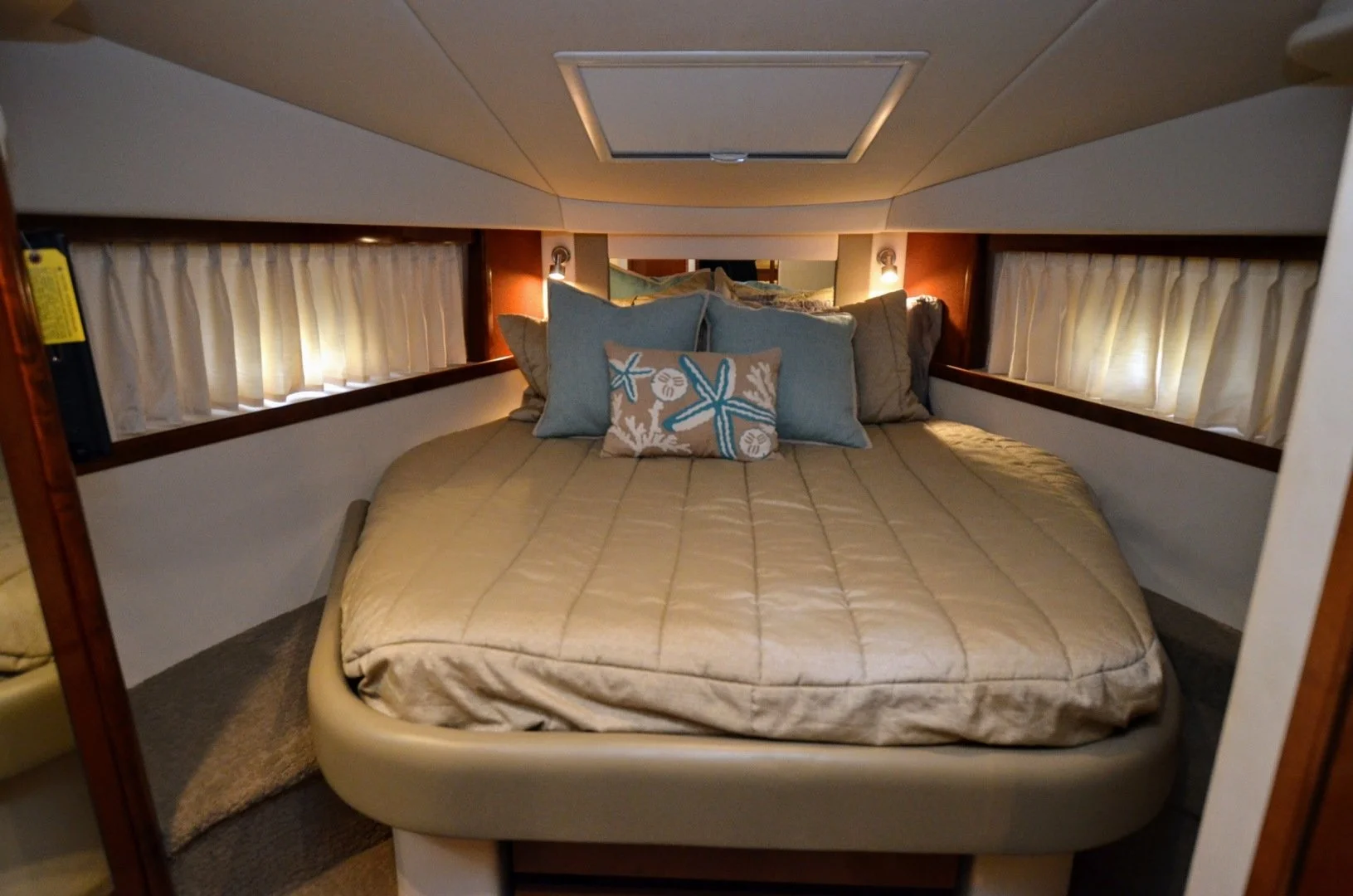 A cozy Sea Ray yacht's bedroom with a bed, beige quilted bedding, and decorative pillows featuring sea shells and starfish, flanked by small windows with curtains.