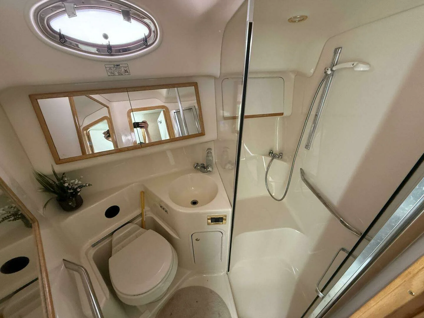 Bathroom aboard Sea Ray yacht with a small shower stall, a toilet, a sink with a mirror above, a small potted plant, and a circular porthole window.