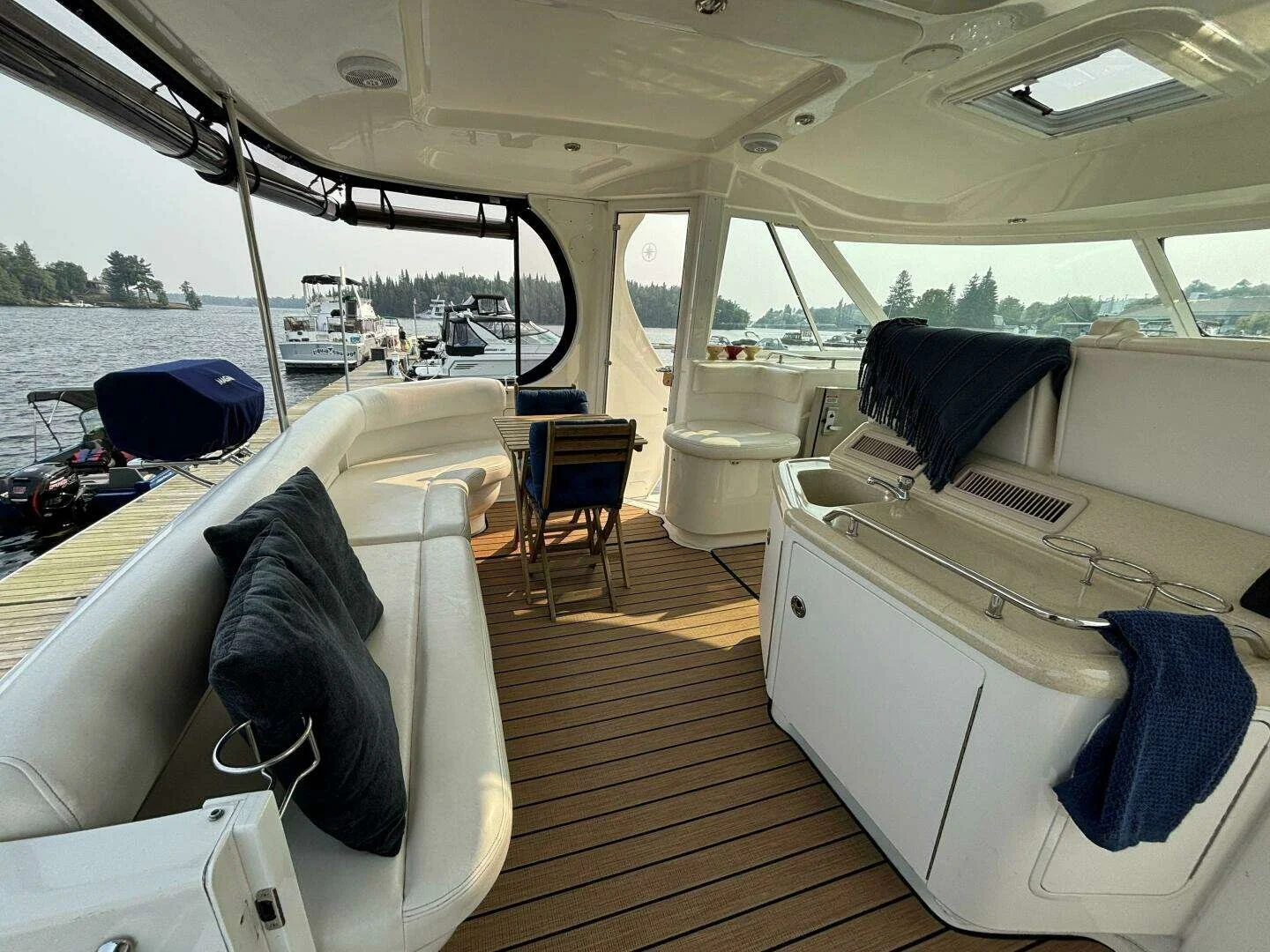 Cockpit of a Sea Ray boat's deck with white cushioned seating, small tables, a black towel, a blue towel, and a view of boats docked on the water through large windows.