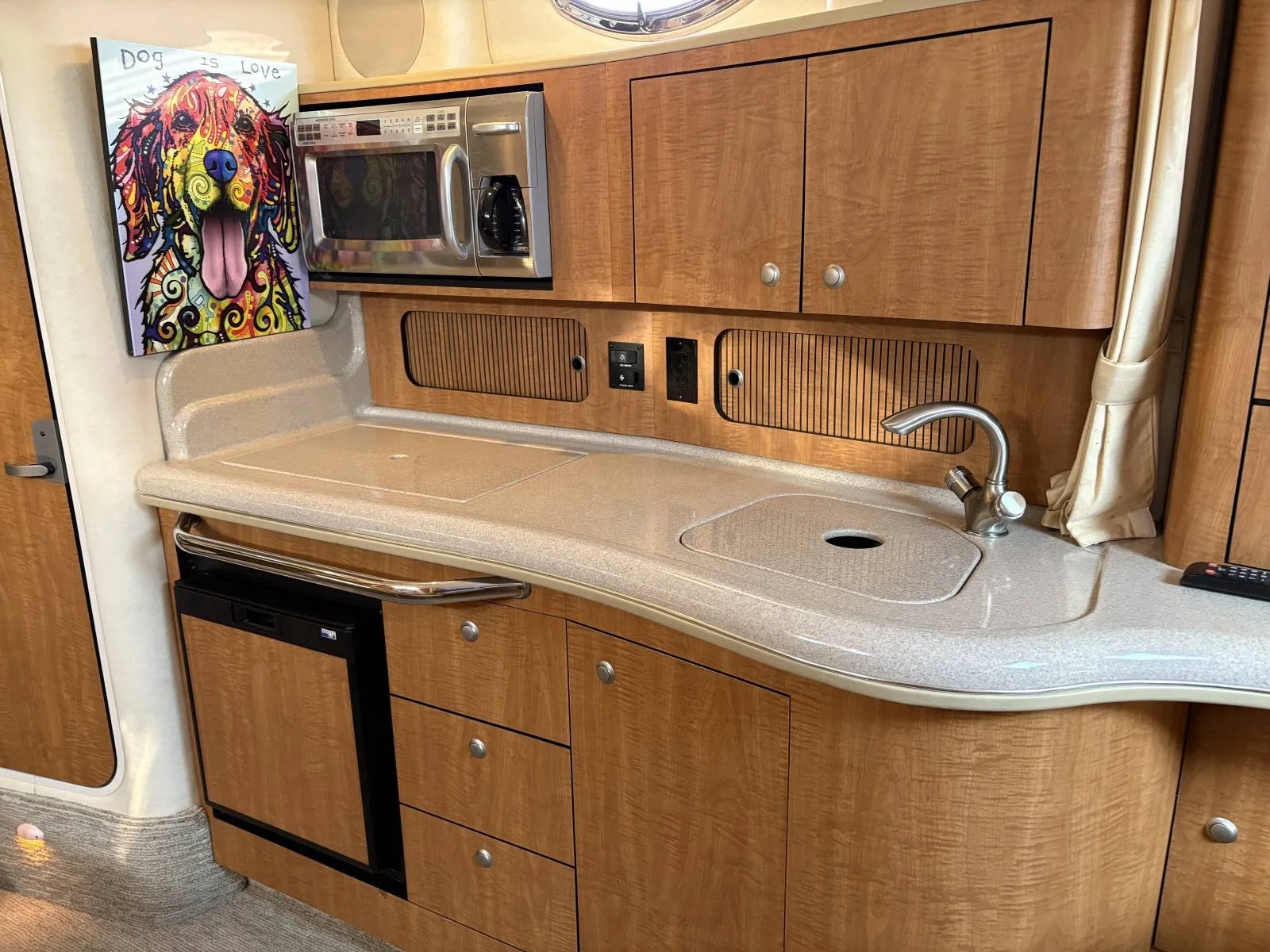 A boat's kitchen area with a beige countertop, stainless steel microwave. There is a small black refrigerator, wooden cabinets, and a beige curtain on the right side.