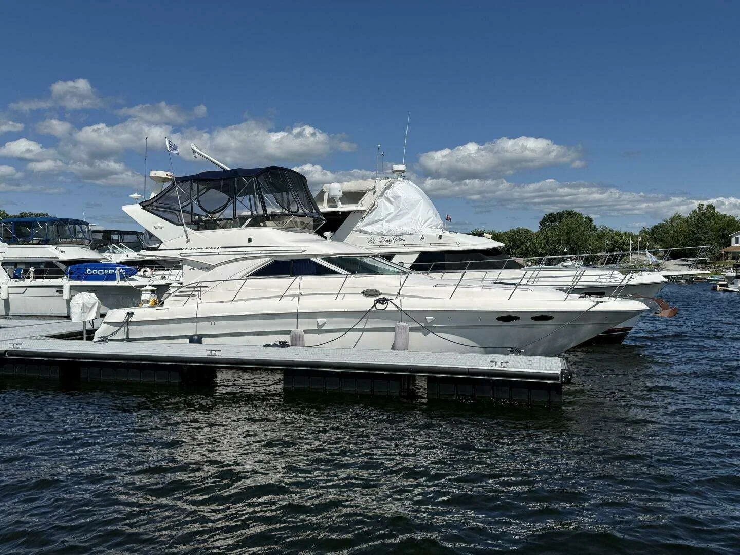 Multiple white yachts docked at a marina on a sunny day with blue sky and scattered clouds. Centered in photo is a 1997 Sea Ray 400 Sedan Bridge boat.
