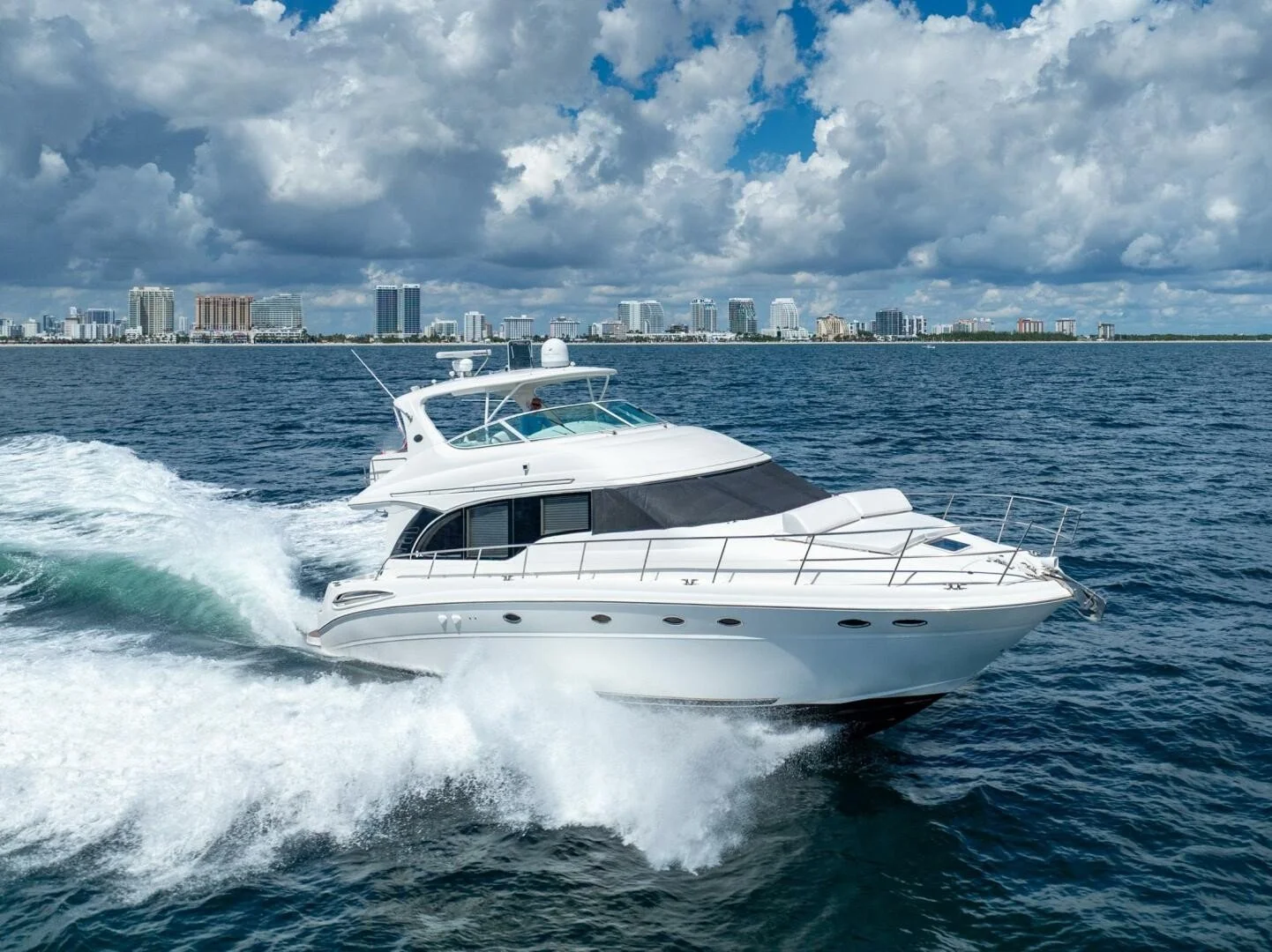 A white luxury yacht sailing on blue waters with a city skyline and cloudy sky in the background. Boat is a Sea Ray 540 Cockpit Motor Yacht.