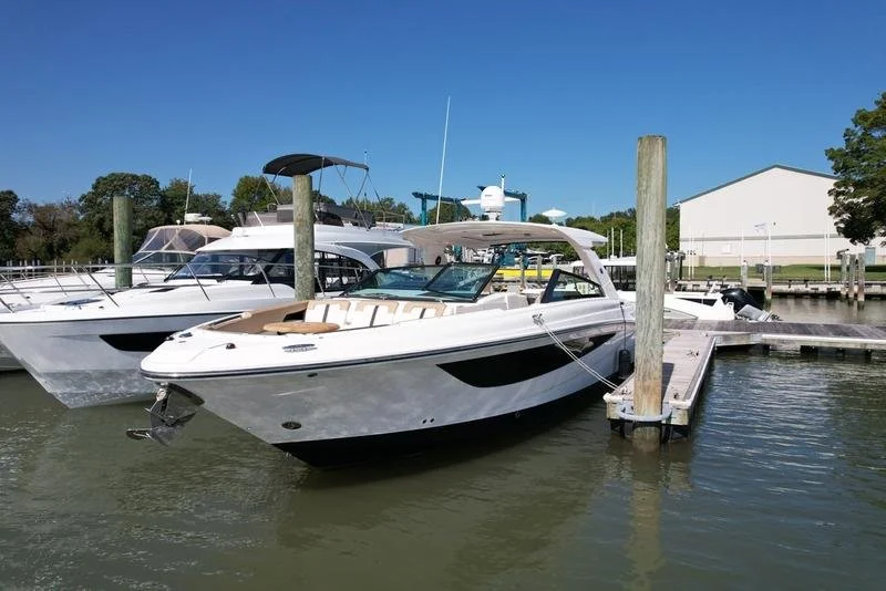 A white motor yacht docked at a marina with a wooden pier, with other boats and a white warehouse in the background under a clear blue sky. Vessel is Sea Ray 400 SLX, 2019 model year boat.