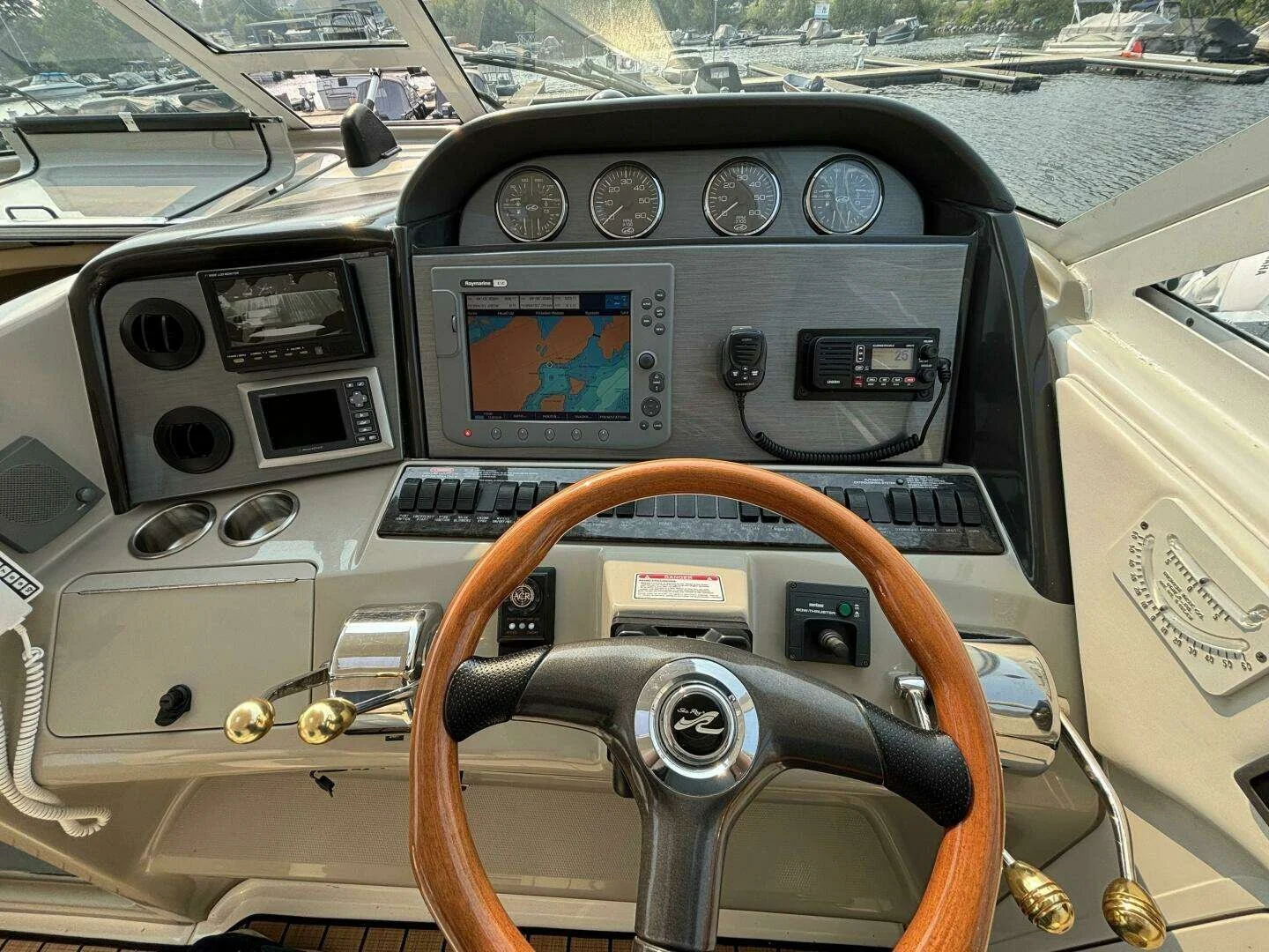 View of a boat's control panel with navigation equipment, gauges, a GPS screen, a radio, and a wooden steering wheel, with marina and boats visible outside the windows. Sea Ray Motor Yacht.