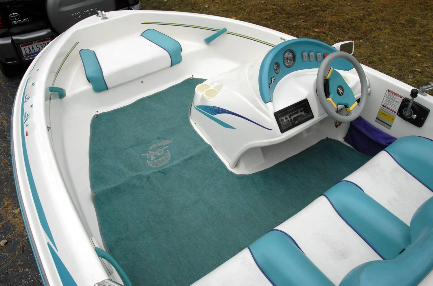 Interior of a small white and blue motorboat with a steering wheel, dashboard gauges, and seating, parked outdoors on grass.