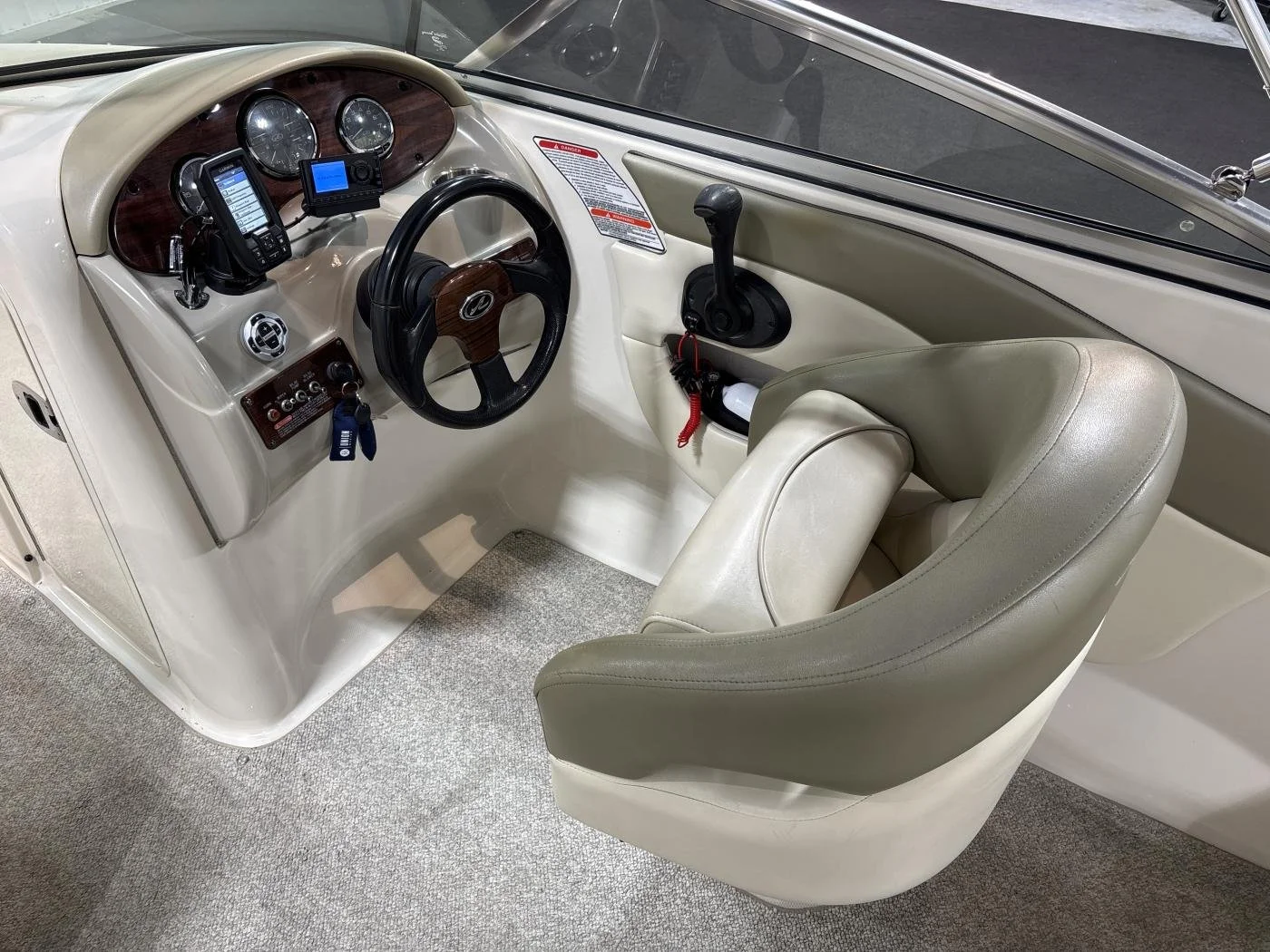 The interior of a boat cabin showing a cream-colored dashboard with gauges, a wireless communication device, and a control panel, alongside a steering wheel, a gear shift lever, and a beige cushioned captain's chair on a carpeted floor.