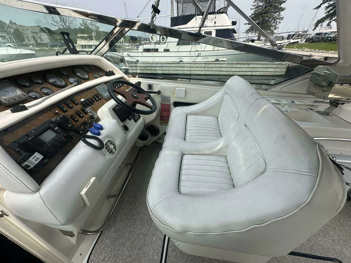 Interior of a boat's cockpit helm area, with a white leather captain's chair, steering wheel, and dashboard with numerous gauges, controls, and communication devices. Through the windshield, other boats are docked at a marina. Sea Ray 380 Sunsport. 
