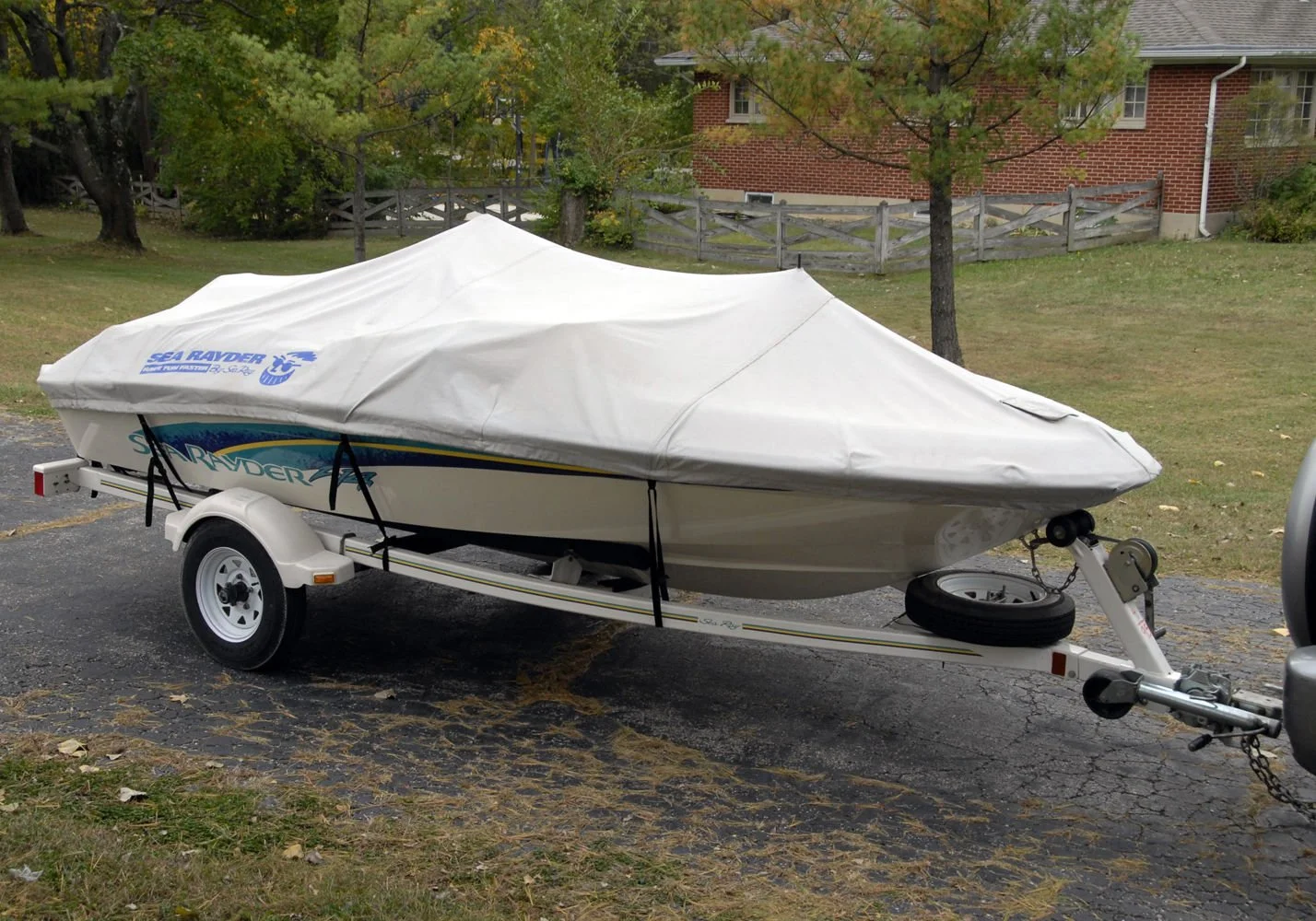 A white motorboat on a trailer with a cover, parked on an asphalt lot near grass and trees.