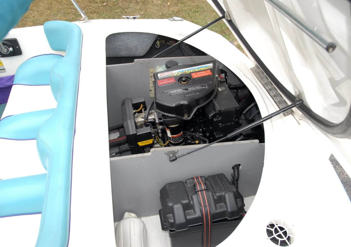 Close-up of a boat's engine compartment with a black engine, blue cushioned seating, and side window, outdoors on grass.
