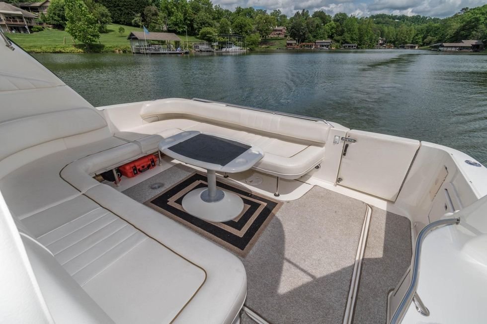 View of the deck on a boat facing a lake with houses and trees in the background, featuring cushioned seating and a small table.