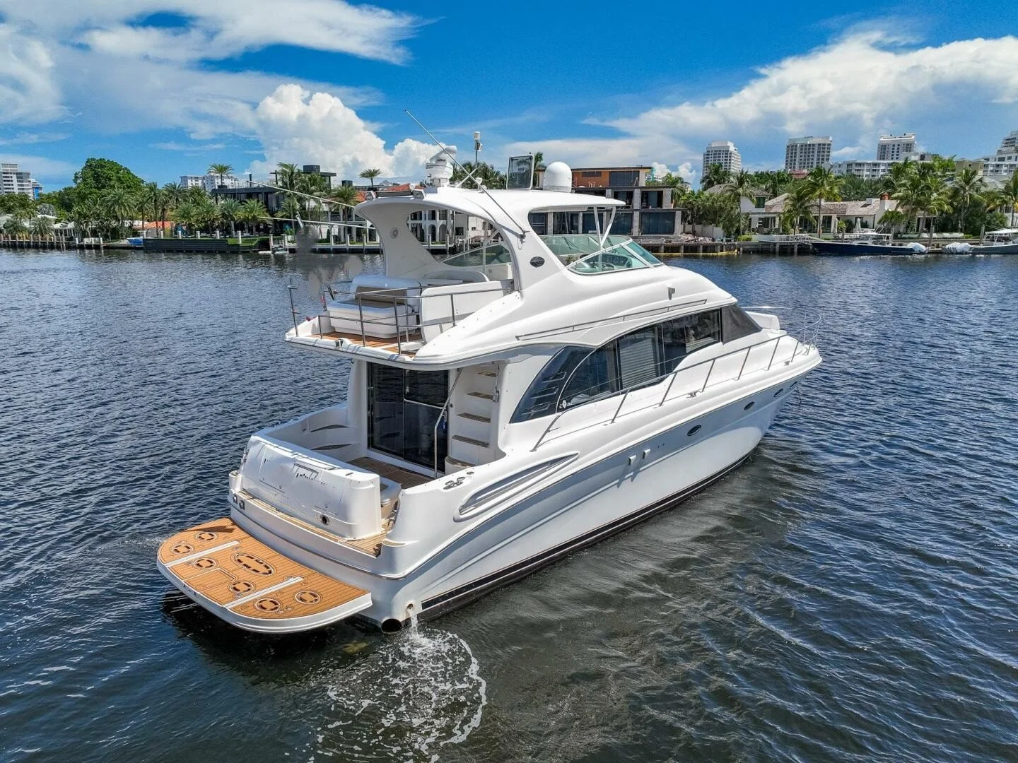 A white luxury yacht on calm water with a modern waterfront neighborhood in the background, palm trees, and a partly cloudy sky.