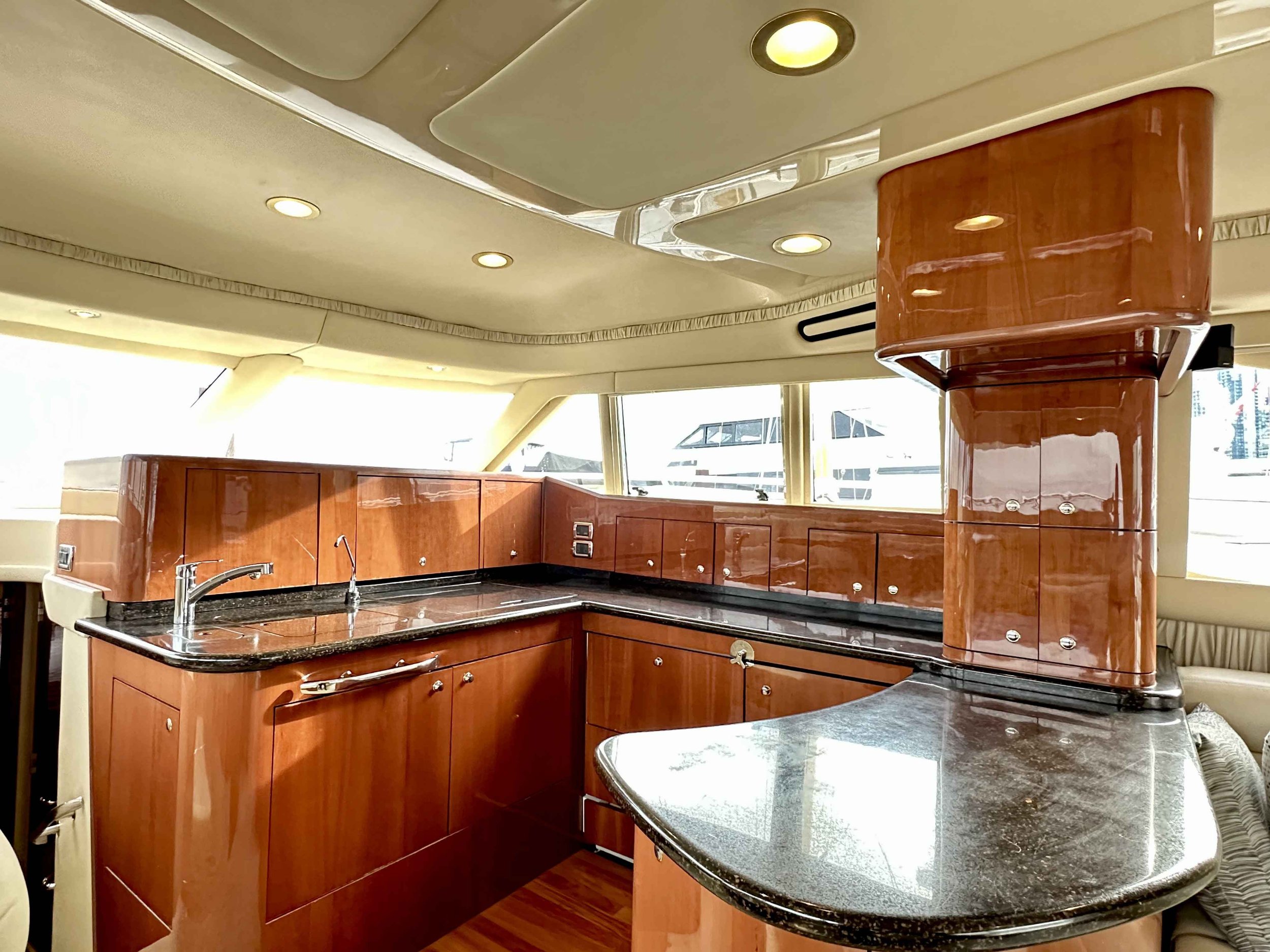Interior view of a yacht galley kitchen with wooden cabinetry, black granite countertops, and a stainless steel faucet, with large windows and a boat dock visible outside.