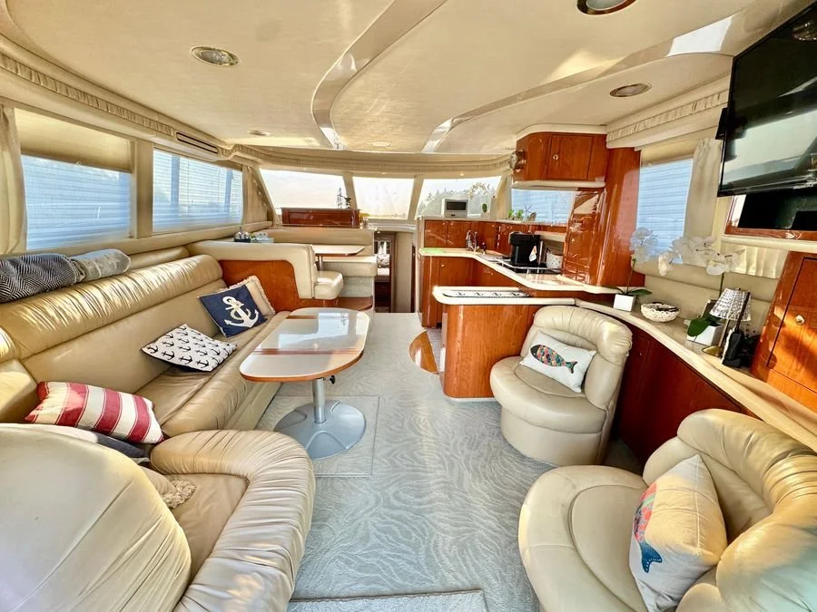 Interior of a yacht or boat with cream leather seating, wooden cabinets, a small table, and decorative pillows, windows with blinds, and a flat-screen TV. Sea Ray 480 Sedan Bridge salon area.