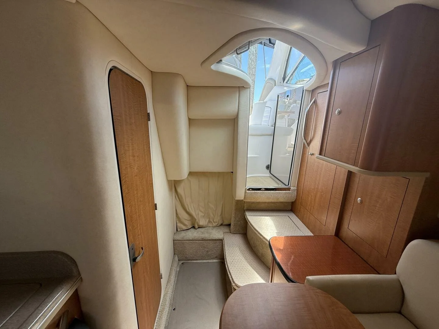 Interior of a compact boat cabin with beige and wood furnishings, a window with a view outside, and a door leading to the exterior deck. Sea Ray Sundancer.