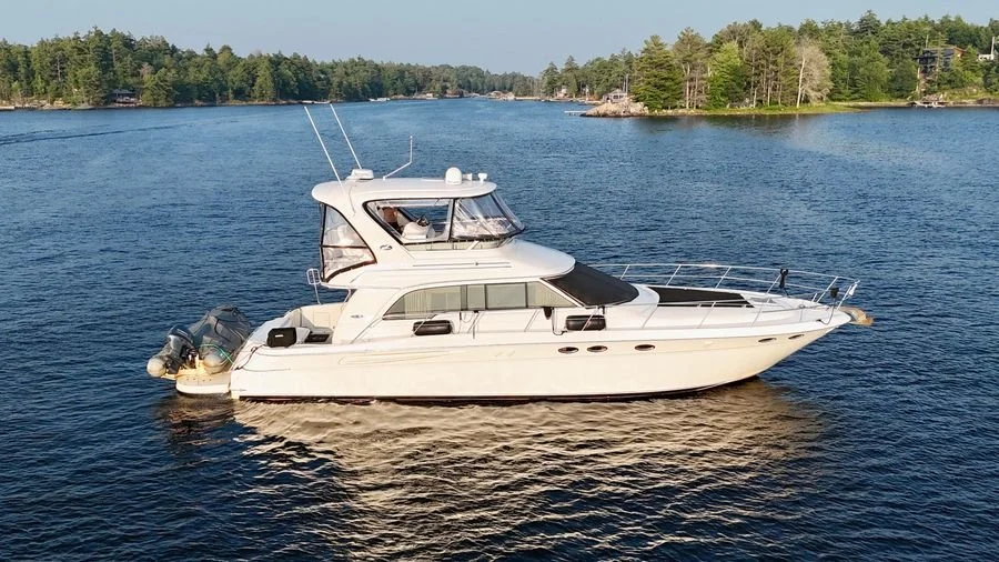 A white yacht with a cabin and flybridge floating on a body of water, with trees and summer cottages in the background. Classic Sea Ray 480 Sedan Bridge boat.