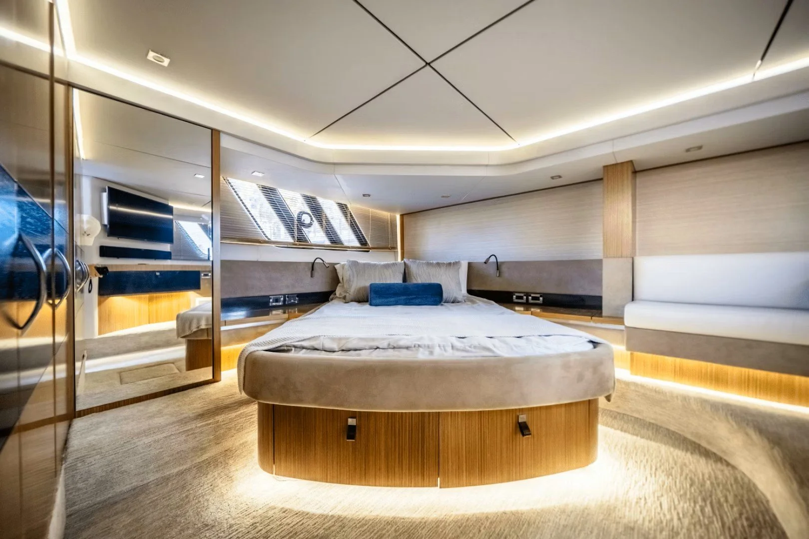 A luxury yacht bedroom with a round bed, beige velvet bedding, and a blue pillow. The room has wood and light-colored paneling, a large window with solar blinds, and ambient lighting along the ceiling and under the bed. Sea Ray motor yacht.