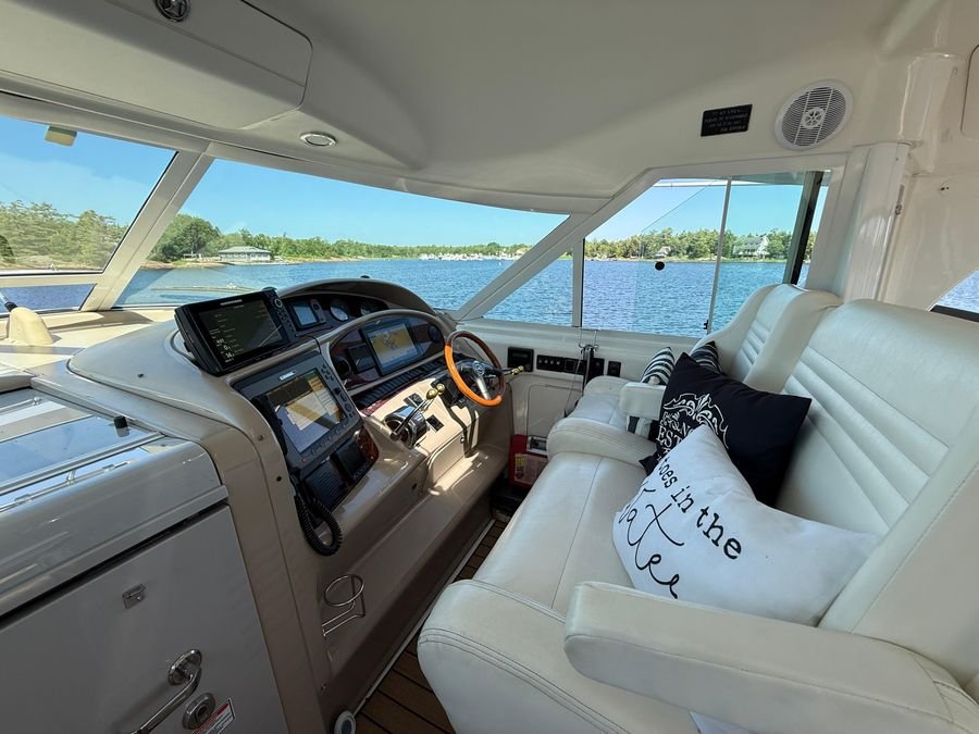 Interior of a yacht with a white leather seating area and navigational electronics, viewing water and greenery through large windows.