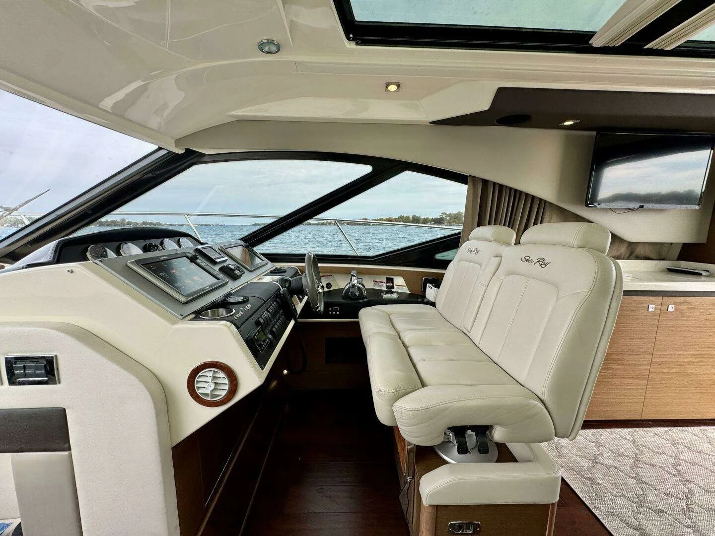 Interior view of a boat's helm station with a white leather captain's chair, navigation dashboard with various controls and screens, a mounted television, and windows showing water and sky outside.