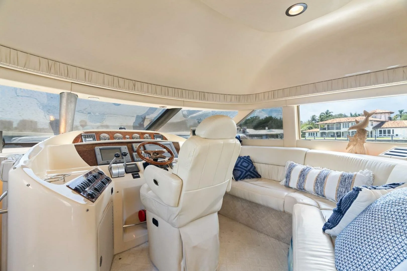 Interior of a yacht with cream leather seating, a wooden steering wheel, and navigation controls, with large windows showing waterfront houses and blue skies.