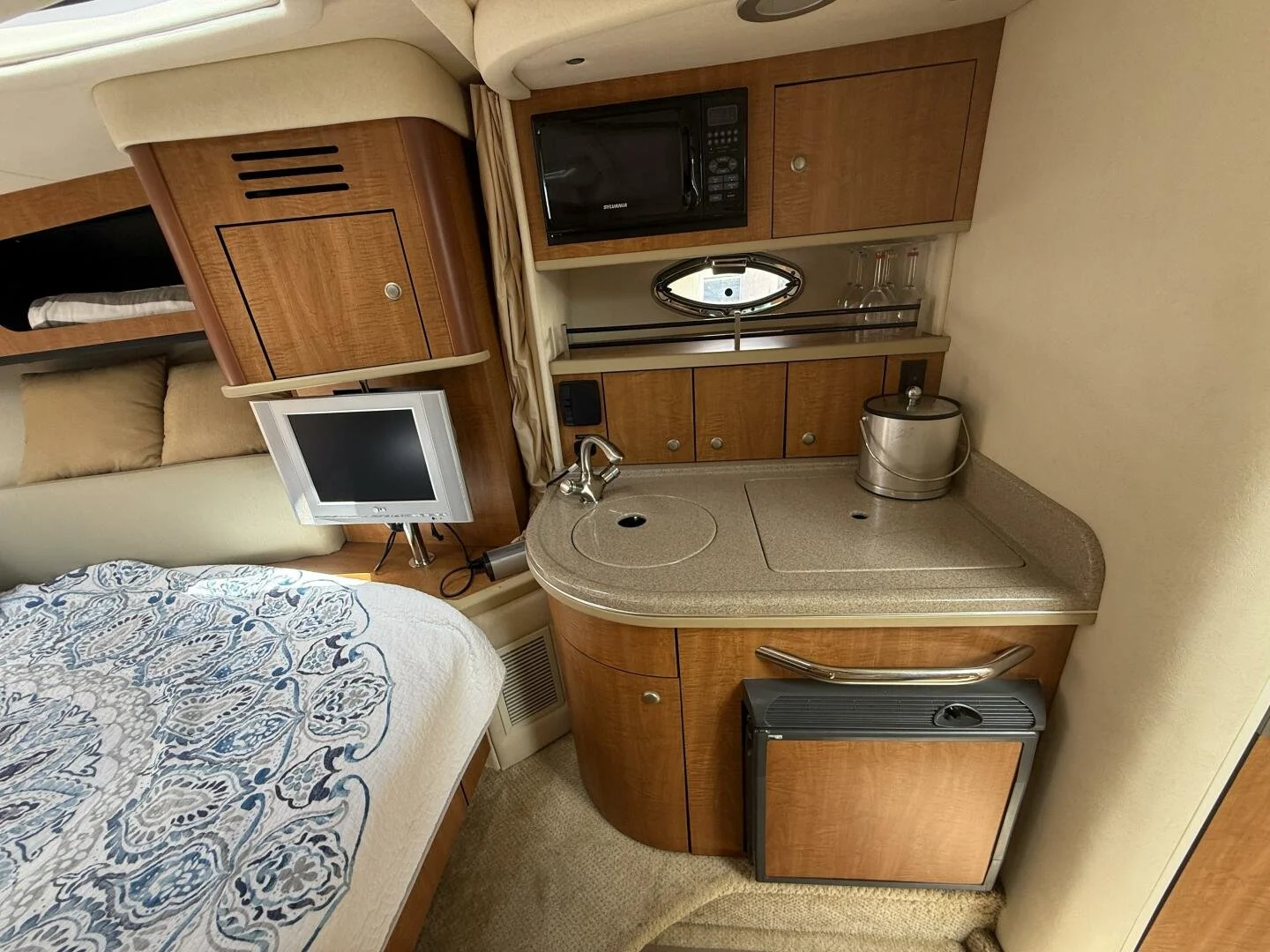 Interior of a Sea Ray boat, featuring a bed with patterned bedding, a small TV, a corner kitchenette galley with a sink, microwave, mini fridge, and storage cabinets, and a window with curtains.