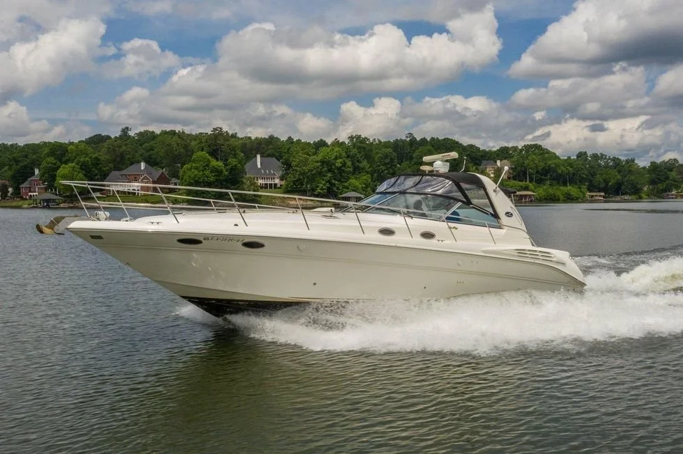 A white yacht boat speeding on a lake with houses and trees in the background under a partly cloudy sky. Express style Sea Ray cruiser, 400 Sundancer.