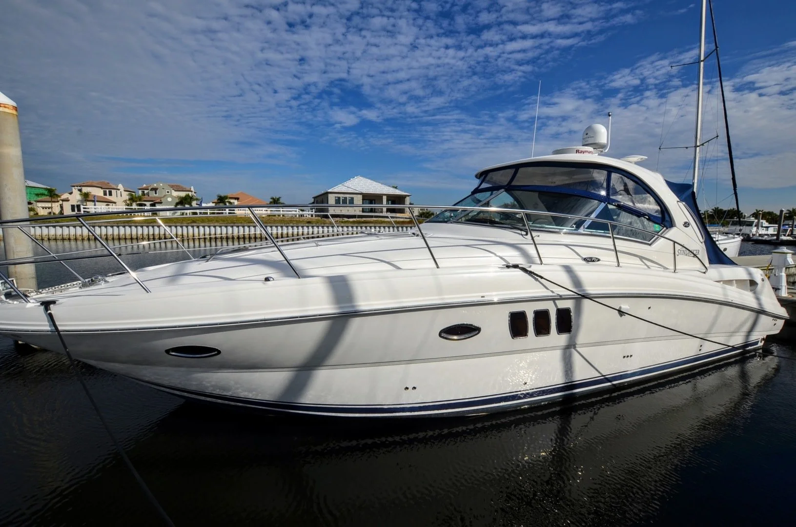 A white yacht docked at a marina with residential houses in the background and a partly cloudy sky overhead. Sea Ray 38 Sundancer ~ 2008