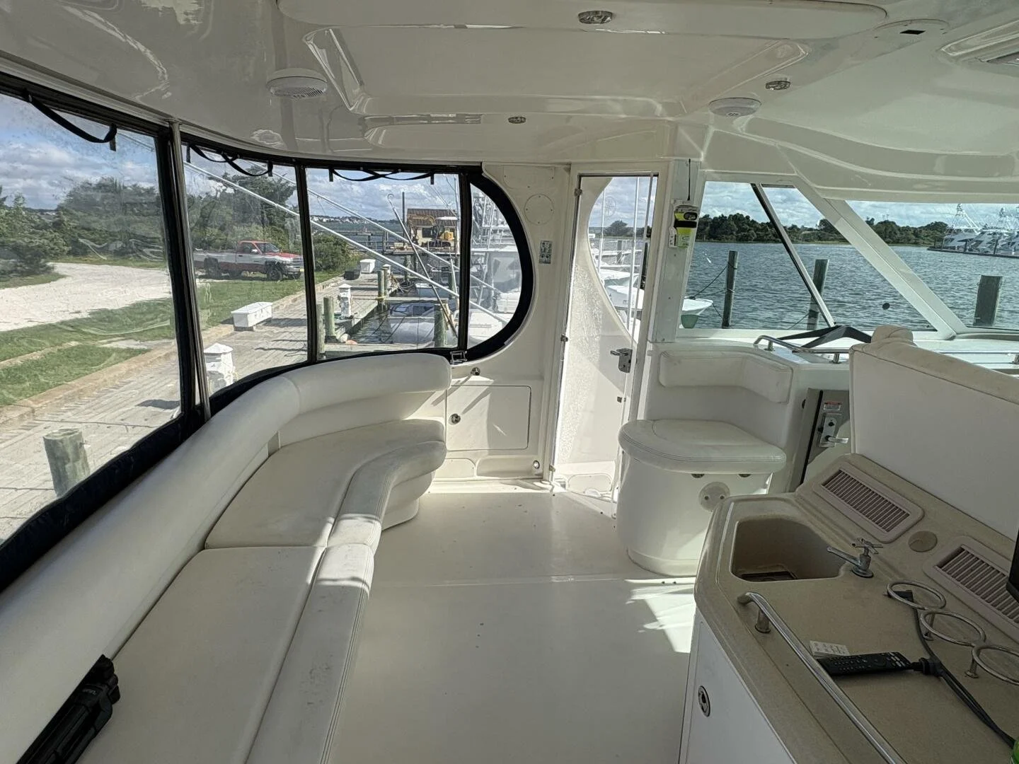 An enclosed cockpit of a Sea Ray yacht, with white seating, a small sink, and a window view of water and other boats docked at a marina.