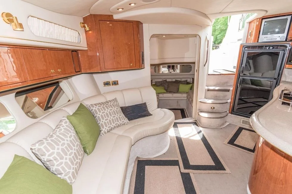 Interior of a luxury yacht with a curved white sofa, green and patterned pillows, wood-paneled cabinets, and a small staircase leading to a sleeping area.