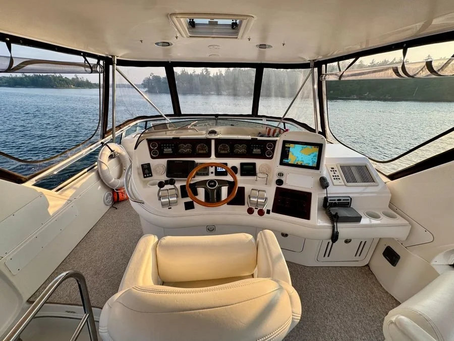 Interior view of a boat cockpit with navigation equipment, steering wheel, and a view of water and shoreline through large front windows. Flybridge on a Sea Ray 480 Sedan Bridge boat.